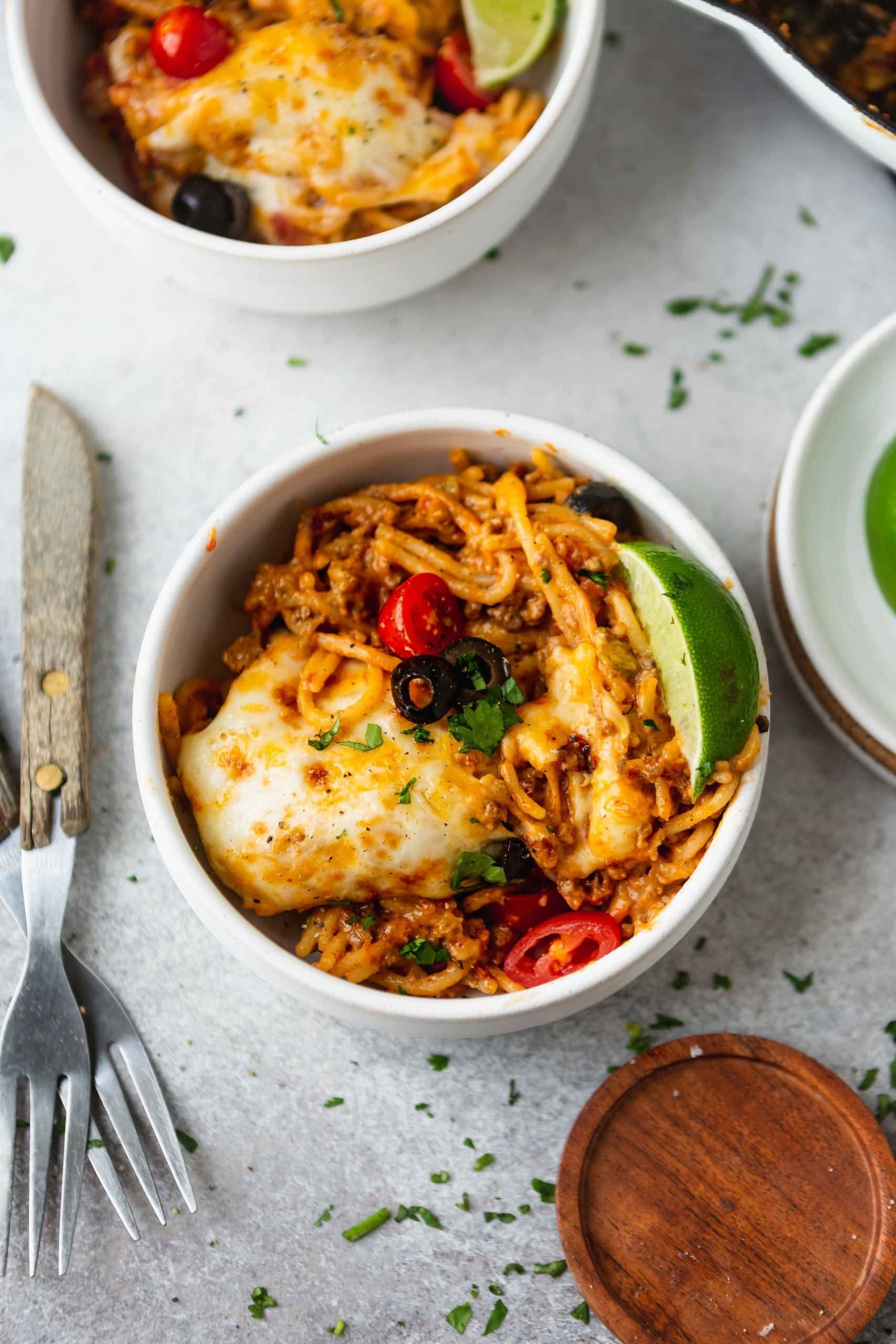 A dinner bowl of cheesy ground beef taco spaghetti. There is a lime wedge on the edge of the bowl. The spaghetti is topped with melted cheese, ground beef, green onions, olives and tomatoes. Two forks are next to the bowl.