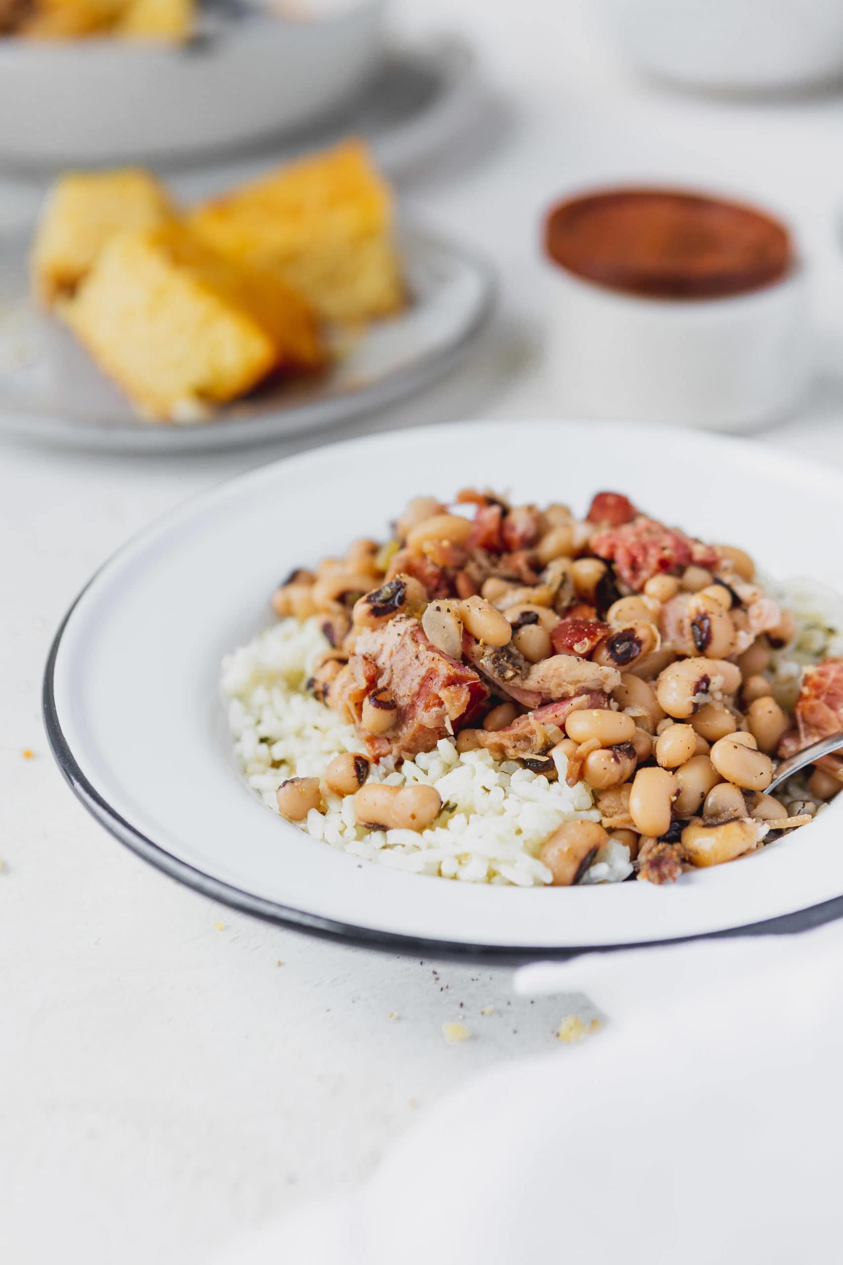 A plate of southern instant pot black eyed peas over rice. There are chunks of ham in the peas and a plate of cornbread in the background.