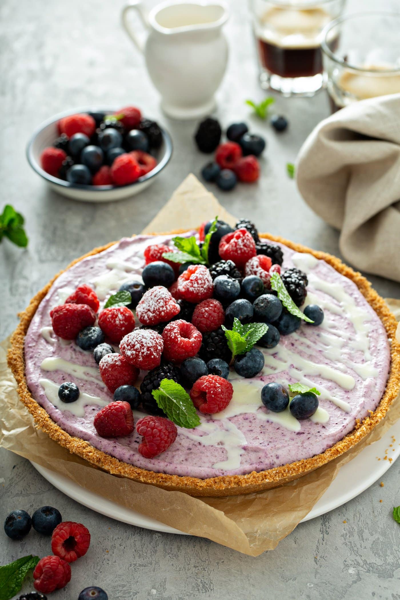 A whole white chocolate berry pie with mixed berries and mint leaves on top. A bowl of mixed berries is in the background and there are berries, mint leaves and a napkin on the table. 