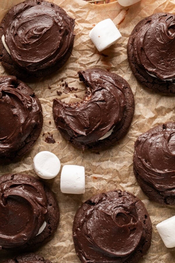 a photo of several chocolate marshmallow cookies with large marshmallows scatted around them sitting on a piece of brown parchment paper