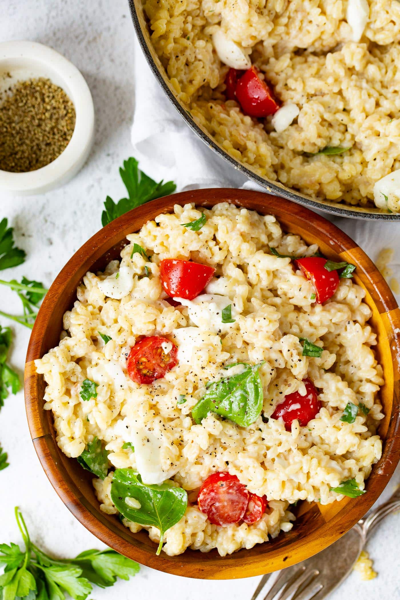 A serving bowl of caprese orzo salad. There are tomatoes, mozzarella chunks and fresh basil leaves in the salad. Another bowl of salad, a container of ground pepper, and a few parsley leaves are on the table next to the bowl.