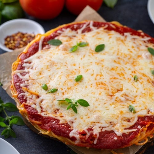A baked spaghetti pie in a pie dish. There are layers of spaghetti, sauce, and melted cheese. Fresh basil is sprinkled on top and three tomatoes, olive oil, a container of dried red pepper flakes, and dinner plates are next to the spaghetti pie.