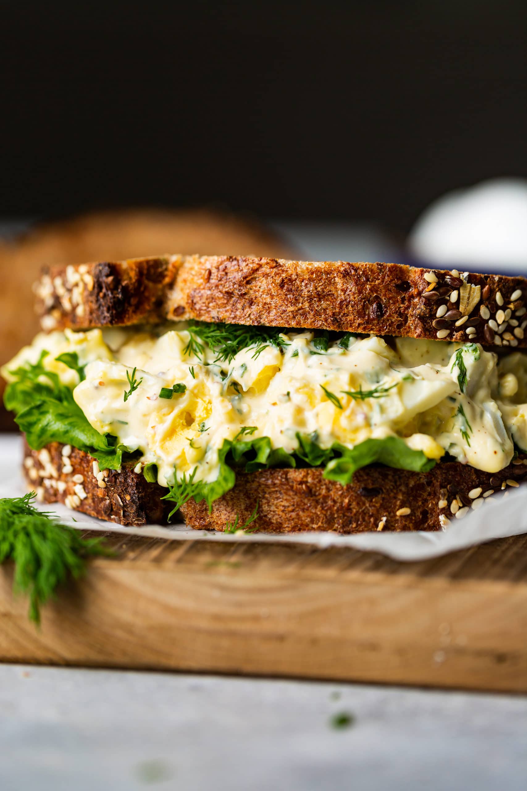 a photo of an egg salad sitting on a piece of parchment paper made on multigrain bread with lettuce and thick layer of egg salad.