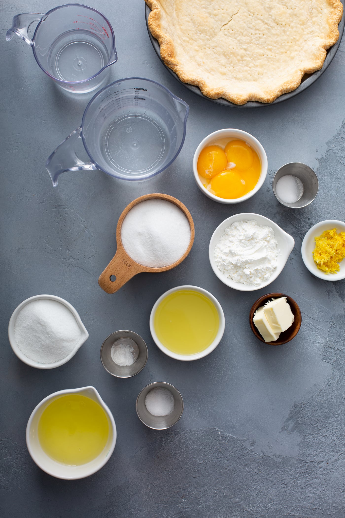 a picture of measuring cups with ingredients for lemon meringue pie. there are containers of eggs, water, sugar, salt lemon juice, lemon zest, butter, etc.