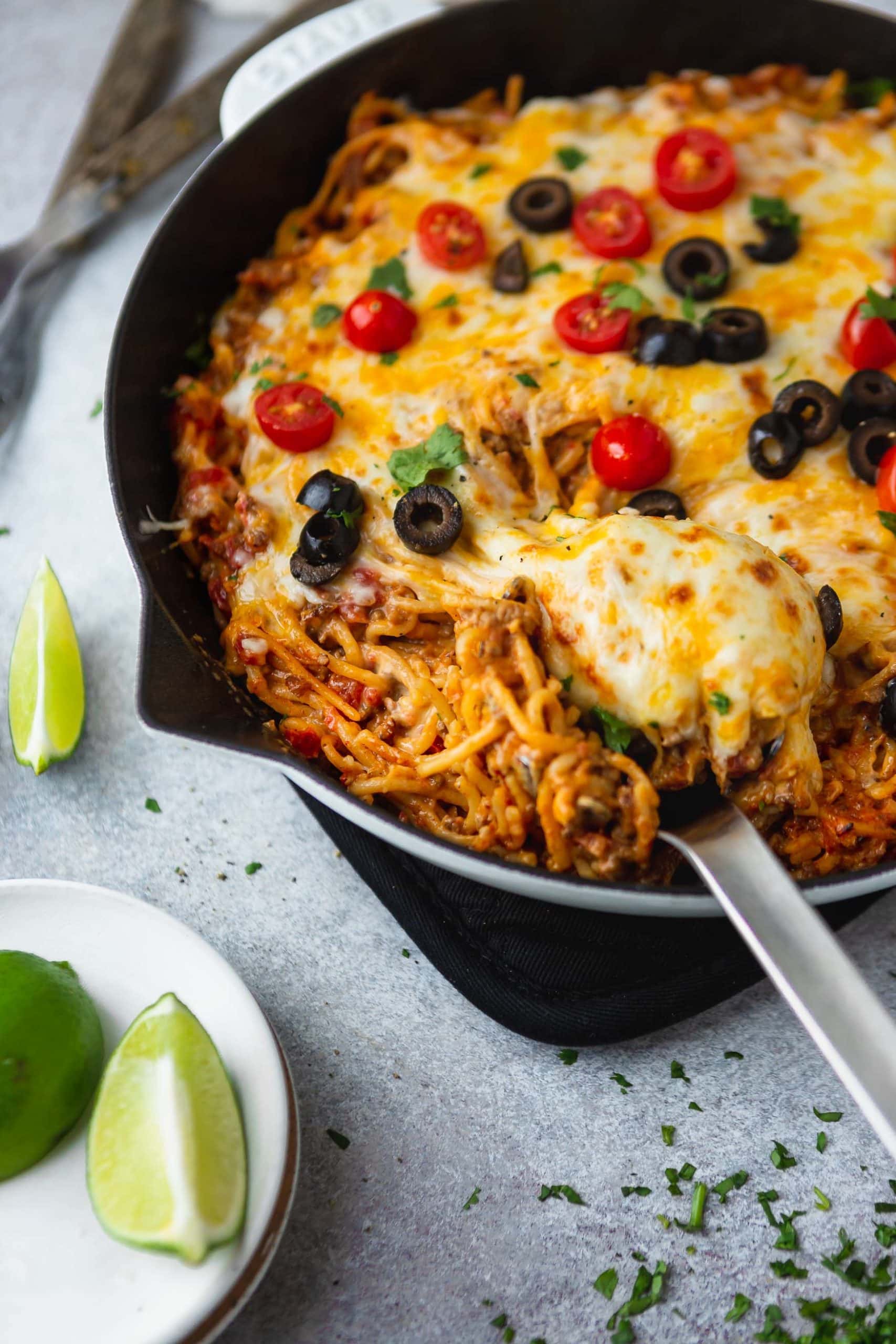 A cast iron skillet filled with cheesy ground beef taco spaghetti. A serving spoon is in the spaghetti and the dish is topped with melted cheese, tomatoes, olives, green onion and cilantro. Lime wedges are on the table next to the skillet.