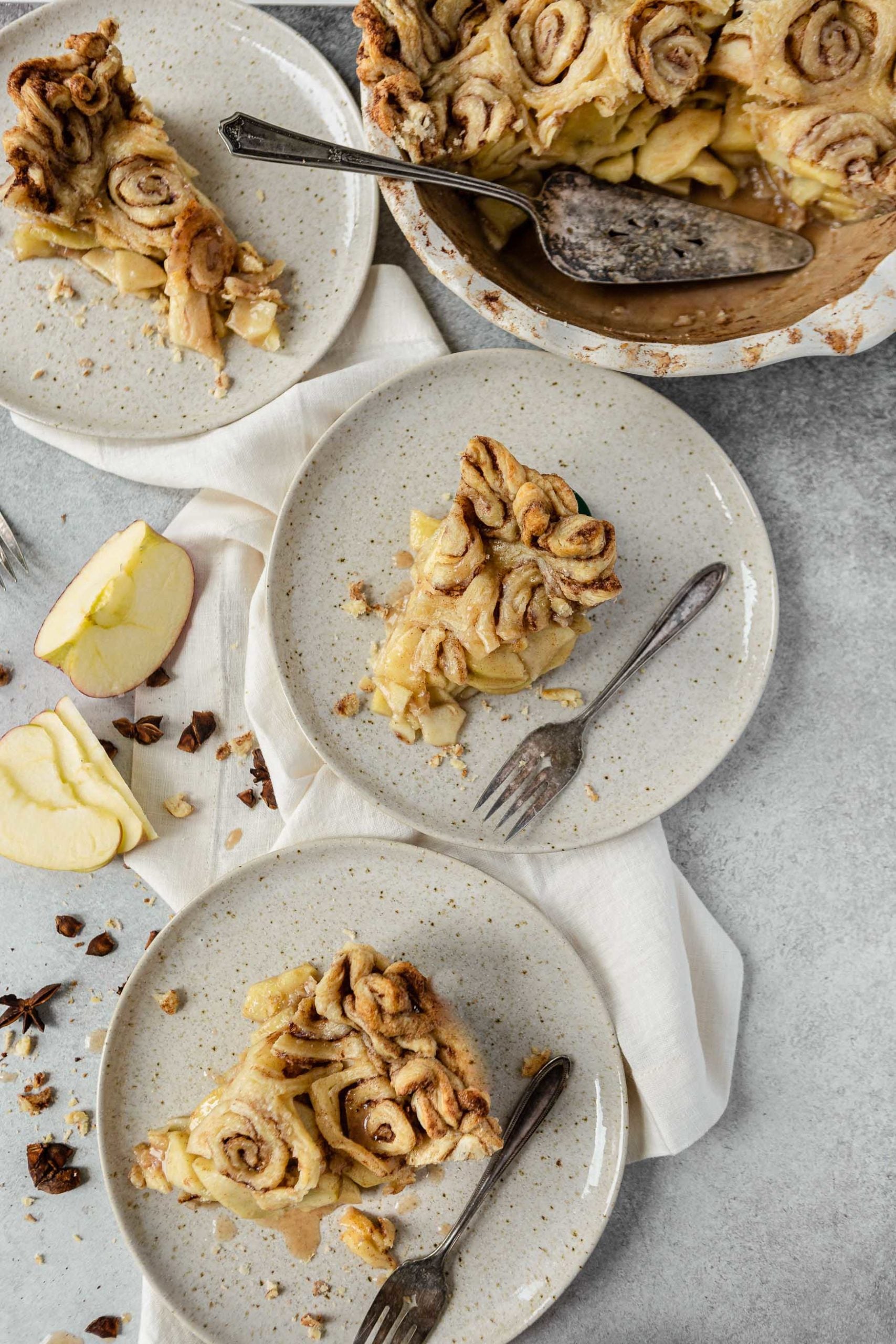 Slices of cinnamon roll apple pie on dessert plates. There are swirled cinnamon rolls as the crust and sliced apples in the background.