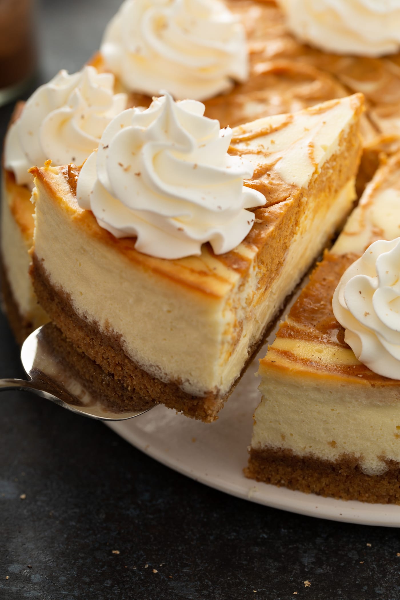 A slice of swirled pumpkin cheesecake being lifted from the whole cheesecake. It has a graham cracker crust and pumpkin filling is swirled into the cheesecake. A dollop of whipped cream is on top.