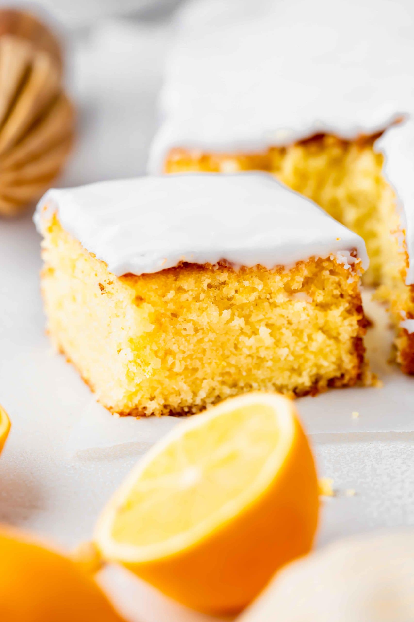 a photo of a piece of lemon cake that has been cut from a square lemon cake. it is covered in a thick white glaze. there is a half of a lemon in front of it.