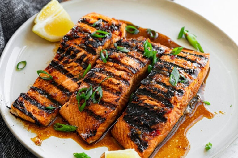 three grilled salmon filets sitting on a white plate sitting in a saucy marinade