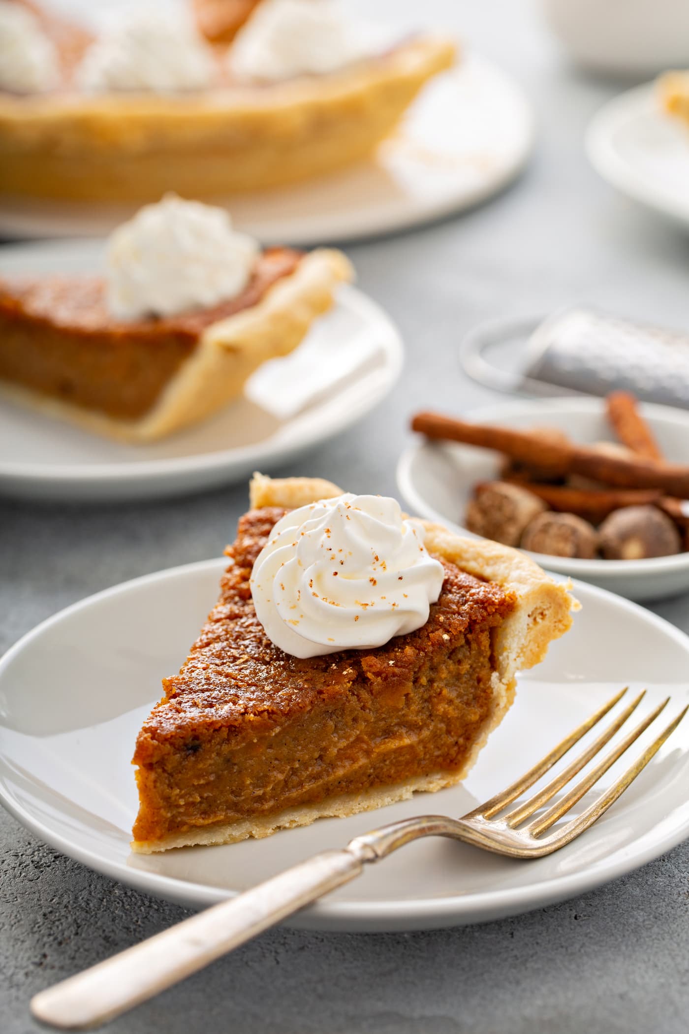 A photo of a slice of sweet potato pie sitting on a white plate with a fork sitting next to it and other servings of pie in the background.