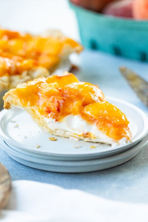 a photo of a single slice of creamy peach pie sitting on a small white dessert plate with a homemade crust, a creamy base layers and topped with a layer of juicy peaches.