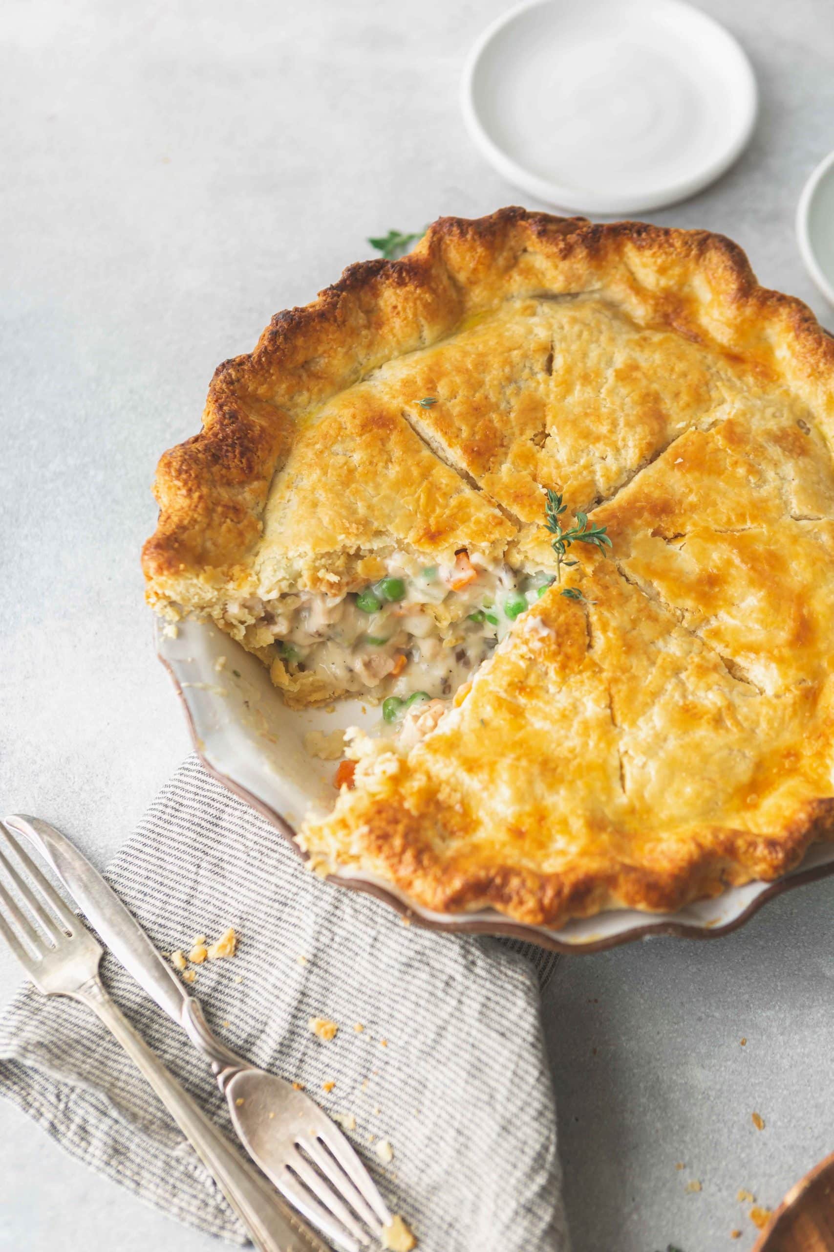 A baked turkey pot pie with a slice removed. The crust is a golden brown and you can see the creamy filling with peas, carrots and turkey. Two forks are next to the pie.