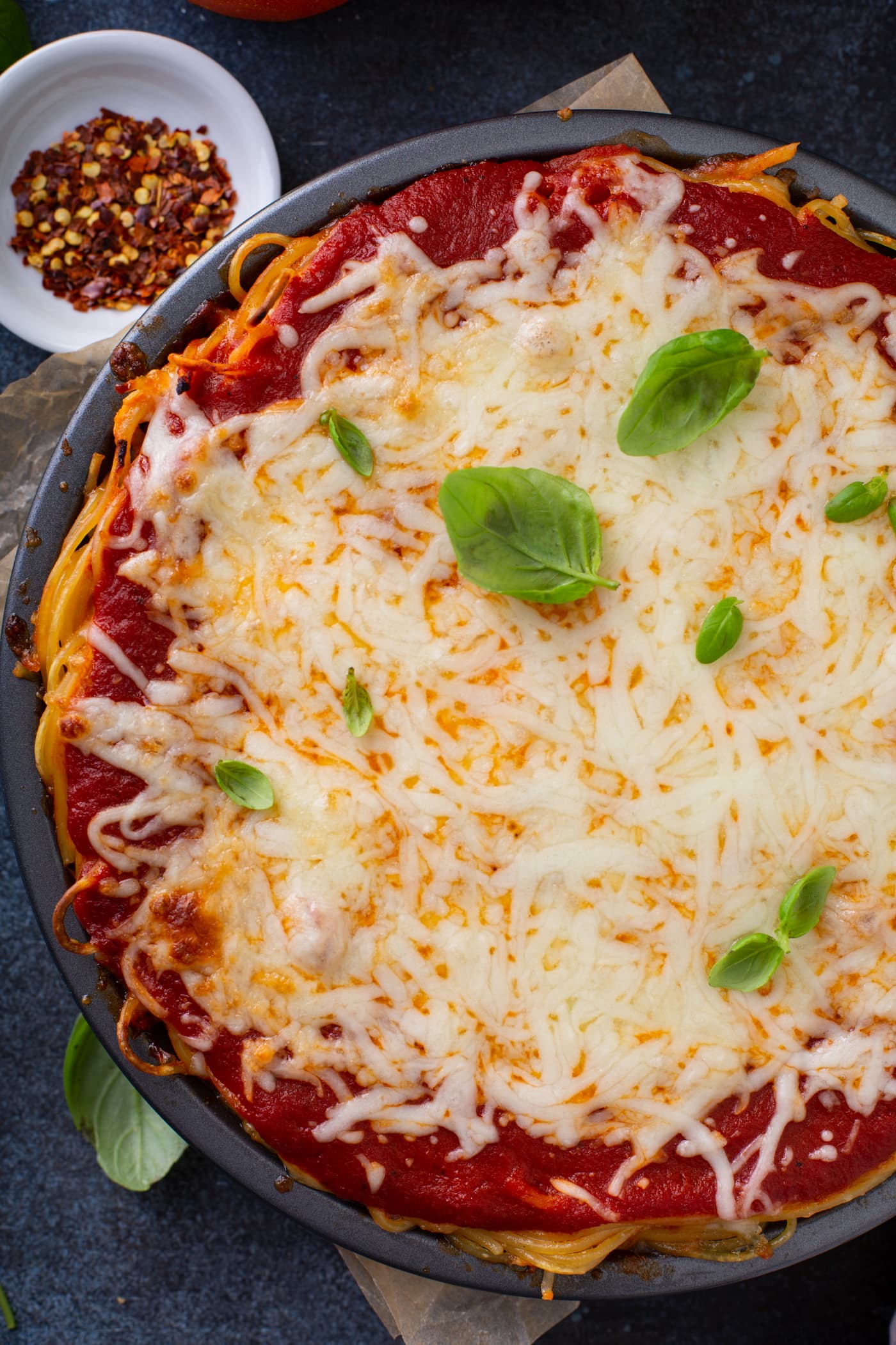 A top view of baked spaghetti pie. There is spaghetti pasta, spaghetti sauce and melted cheeses with a few basil leaves on top. A small bowl of dried pepper flakes is behind the pie.