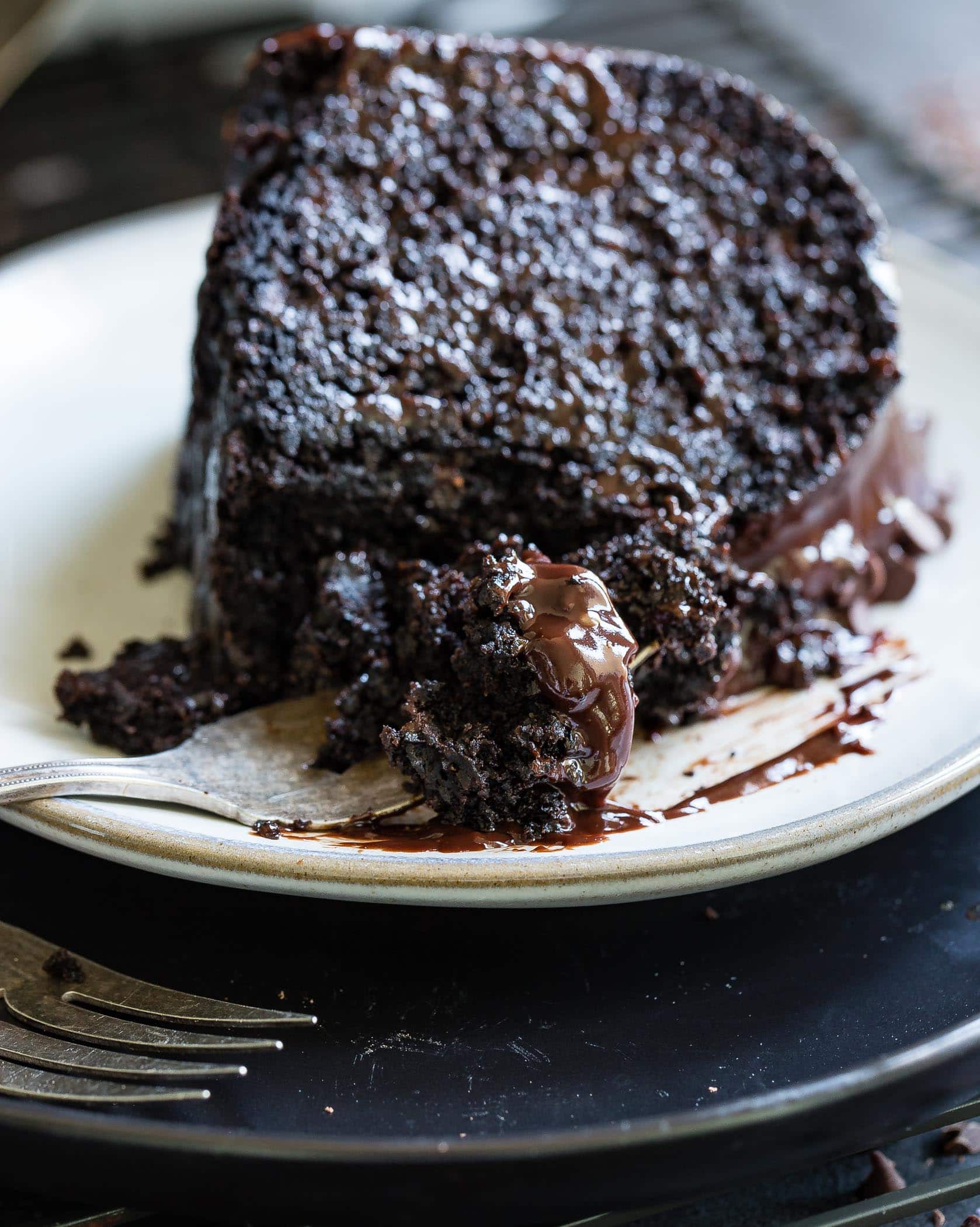 A slice of zucchini chocolate bundt cake on a white dessert plate. The cake is dark, moist chocolate with a chocolate glaze. A fork is beside the plate.