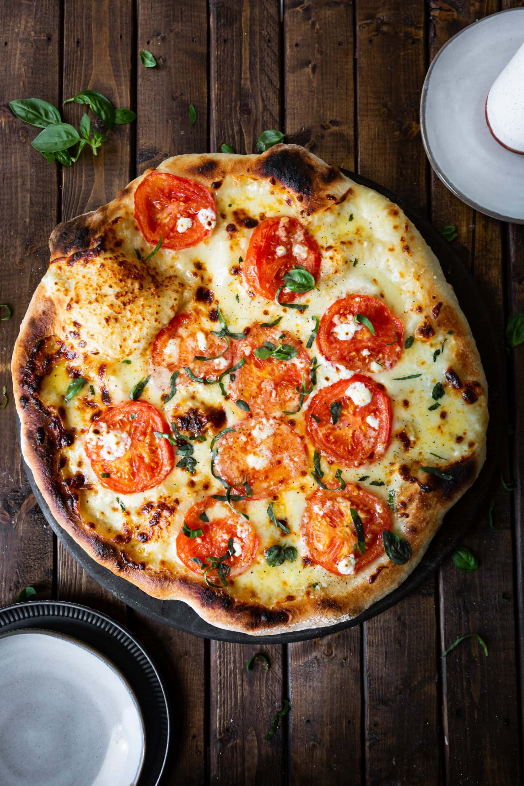 a photo of a whole margherita pizza with golden bubbled crust, slices of tomato, chopped fresh basil and chunks of feta cheese.