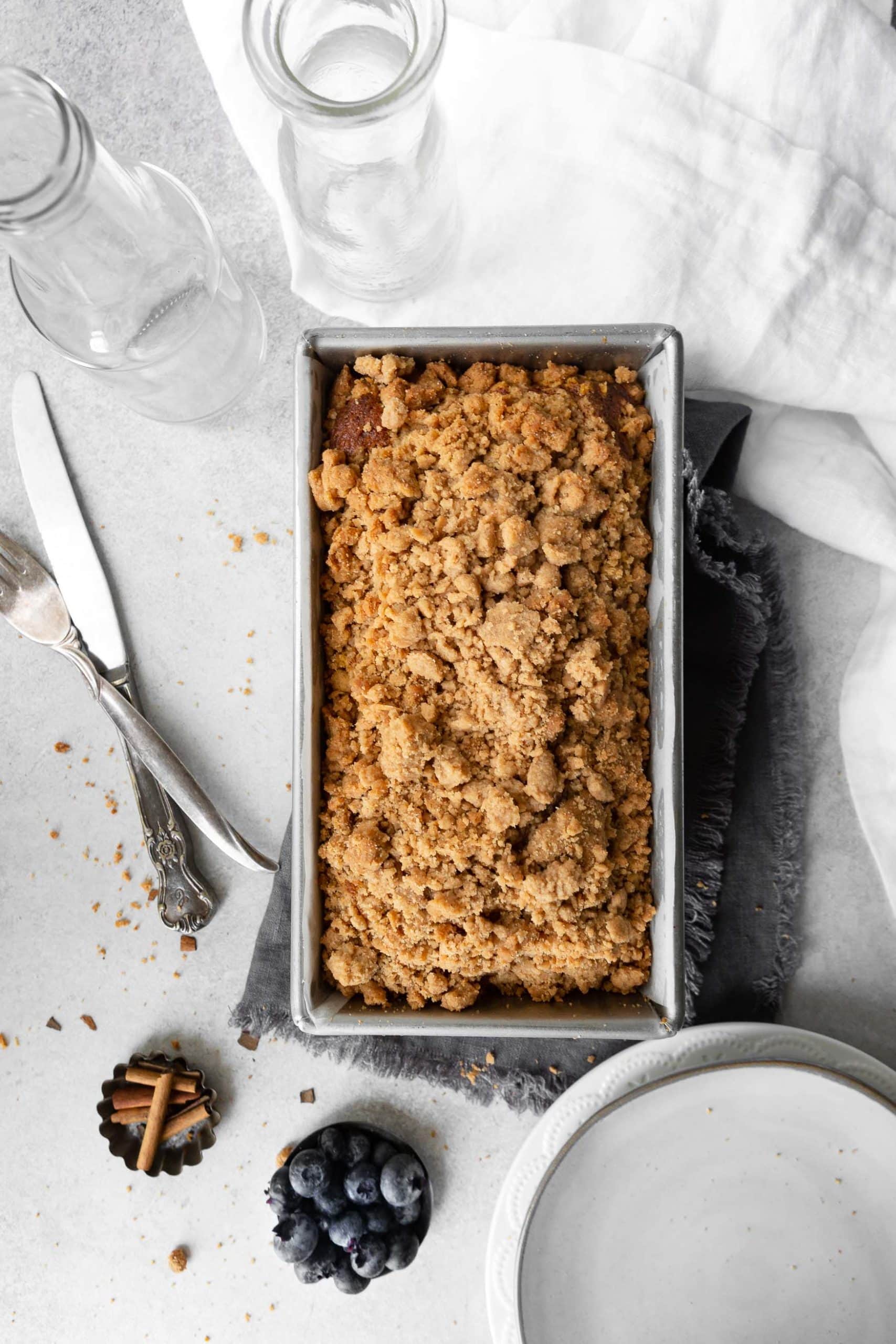 a photo of a loaf of pumpkin bread with cinnamon crumb topping still in the bread pan with a small cup of blueberries sitting next the pan and a small white serving plate.