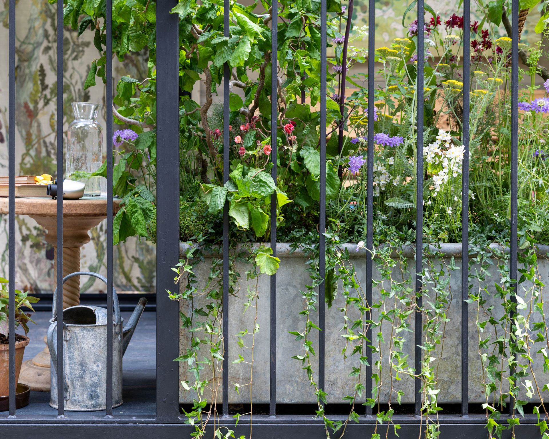 balcony with metal raised bed planted with summer flowers, a watering can and a birdbath