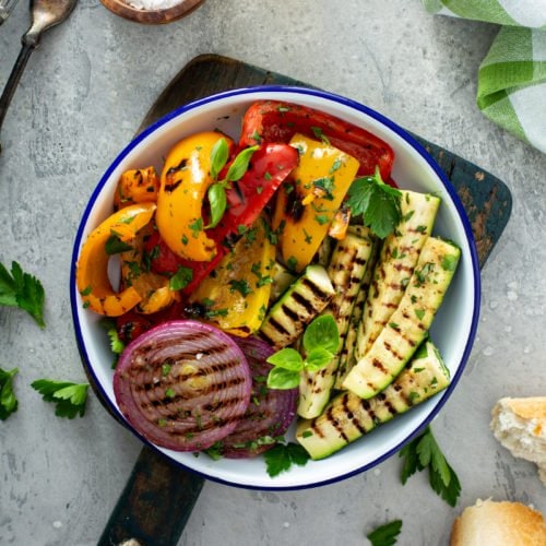 A serving plate of grilled vegetables sitting on a wooden cutting board. The grilled red onion, red and yellow bell peppers, and zucchini are topped with parsley leaves. A few parsley leaves are scattered on the table. A wooden container of salt, two forks and a glass container of oil is also sitting on the table. A broken bread roll is next to the veggies.