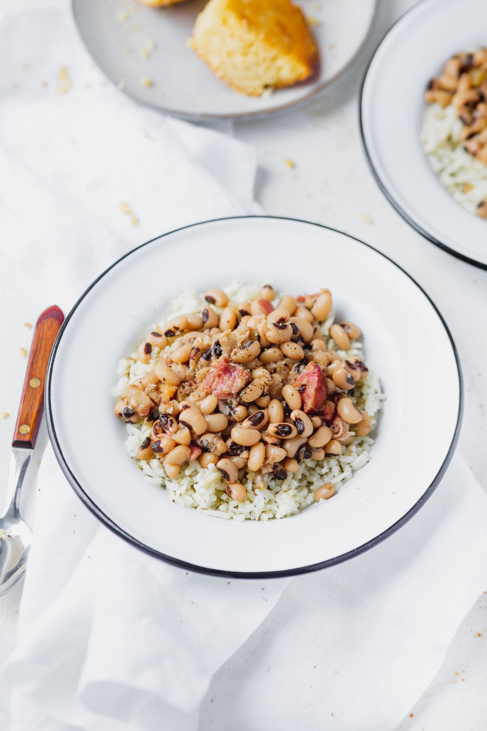 A plate of instant pot black eyed peas over rice. There is a spoon next to the plate and a plate of cornbread in the background.