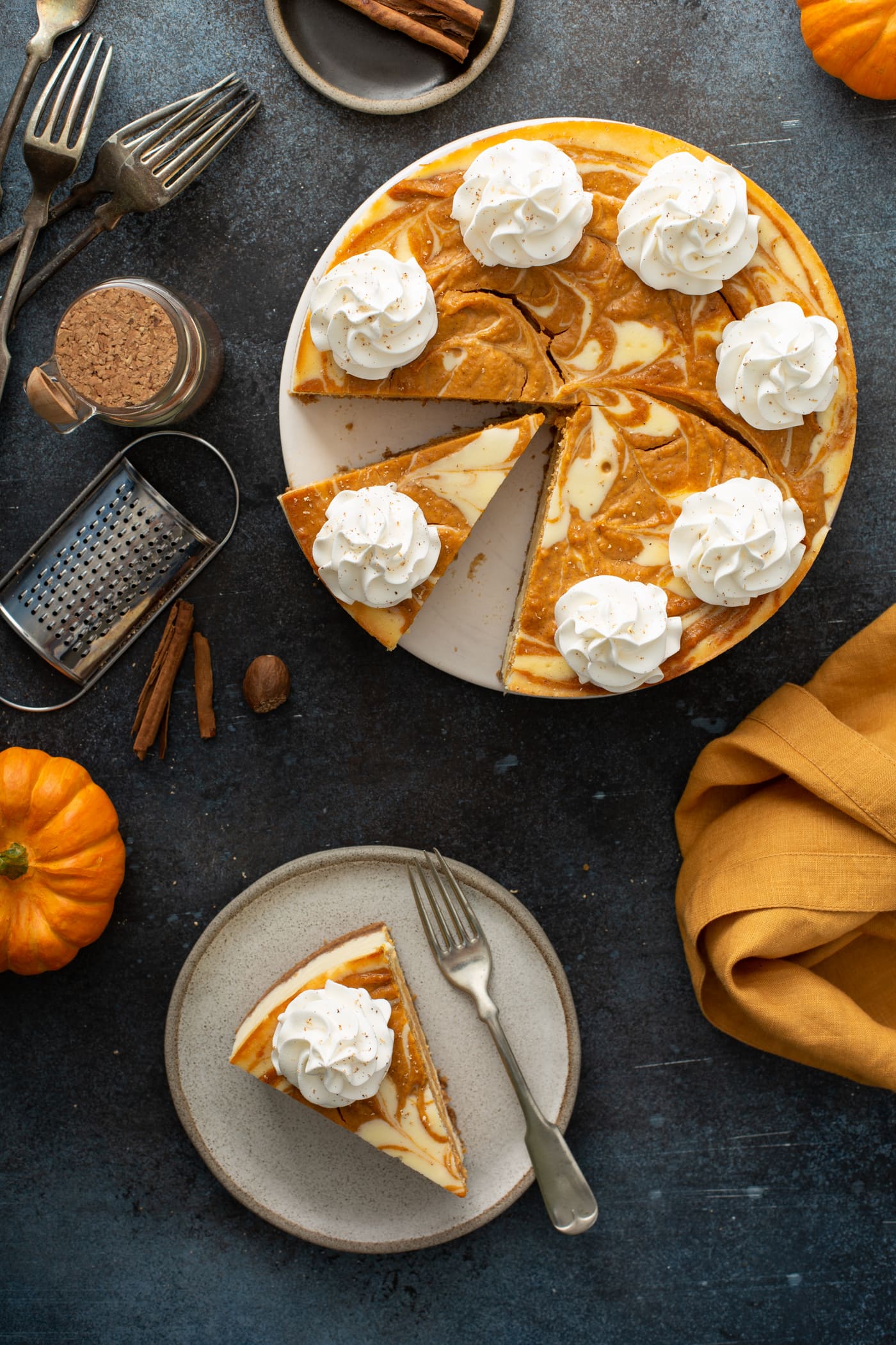 A plate with a slice of swirled pumpkin cheesecake and a fork. The rest of the cheesecake is in the background with cinnamon sticks, a small pumpkin and an orange napkin.
