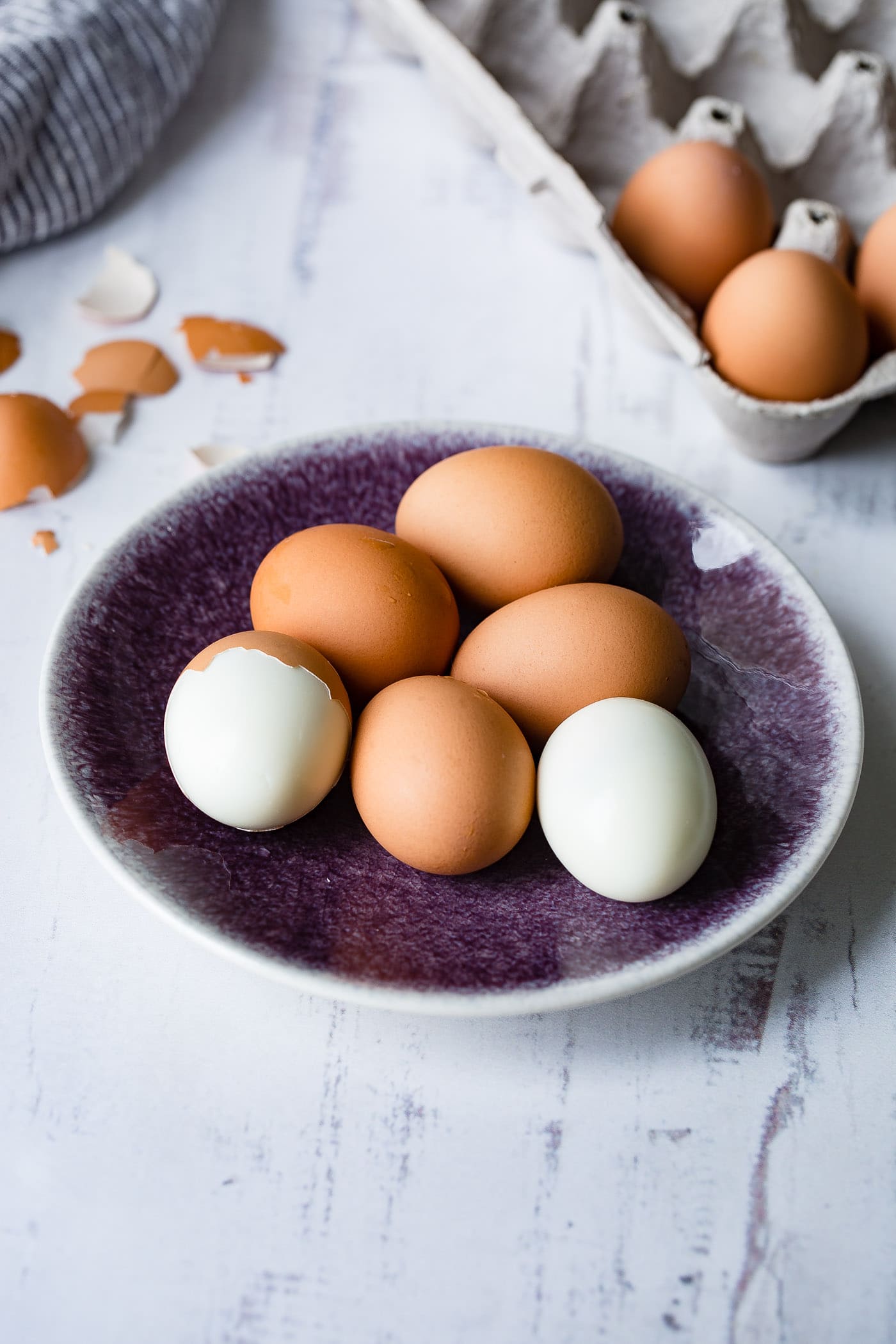 A plate with 6 brown hard boiled eggs. One has been peeled and one is partially peeled. There are brown egg shell and a carton of eggs in the background.