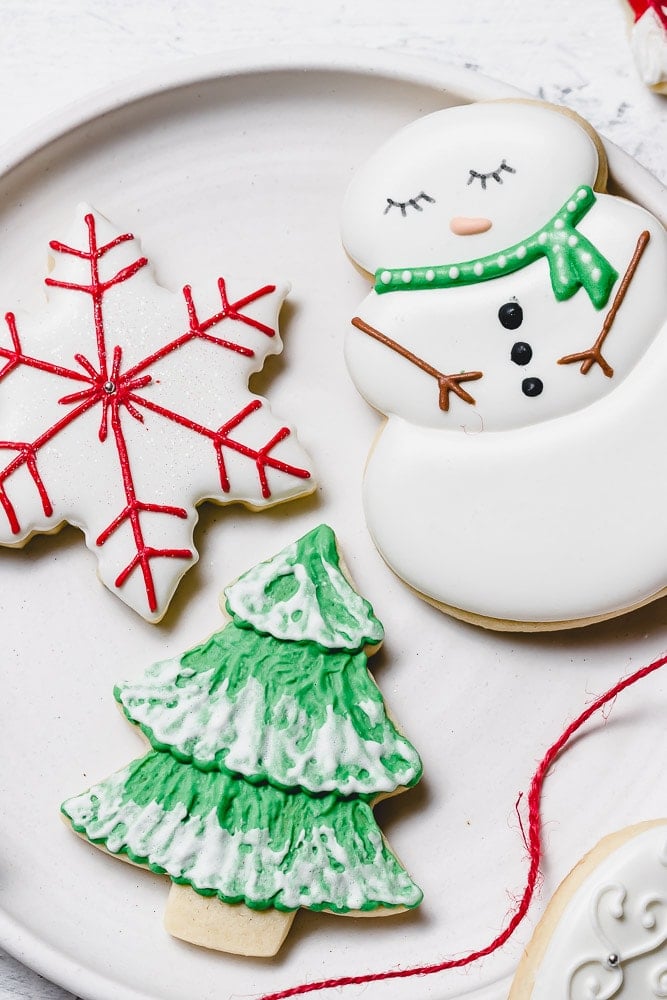 Christmas Sugar cookies with royal icing. There is a snowman with a green scarf, a tree with green icing and white accents like snow, and a white star with red designs.