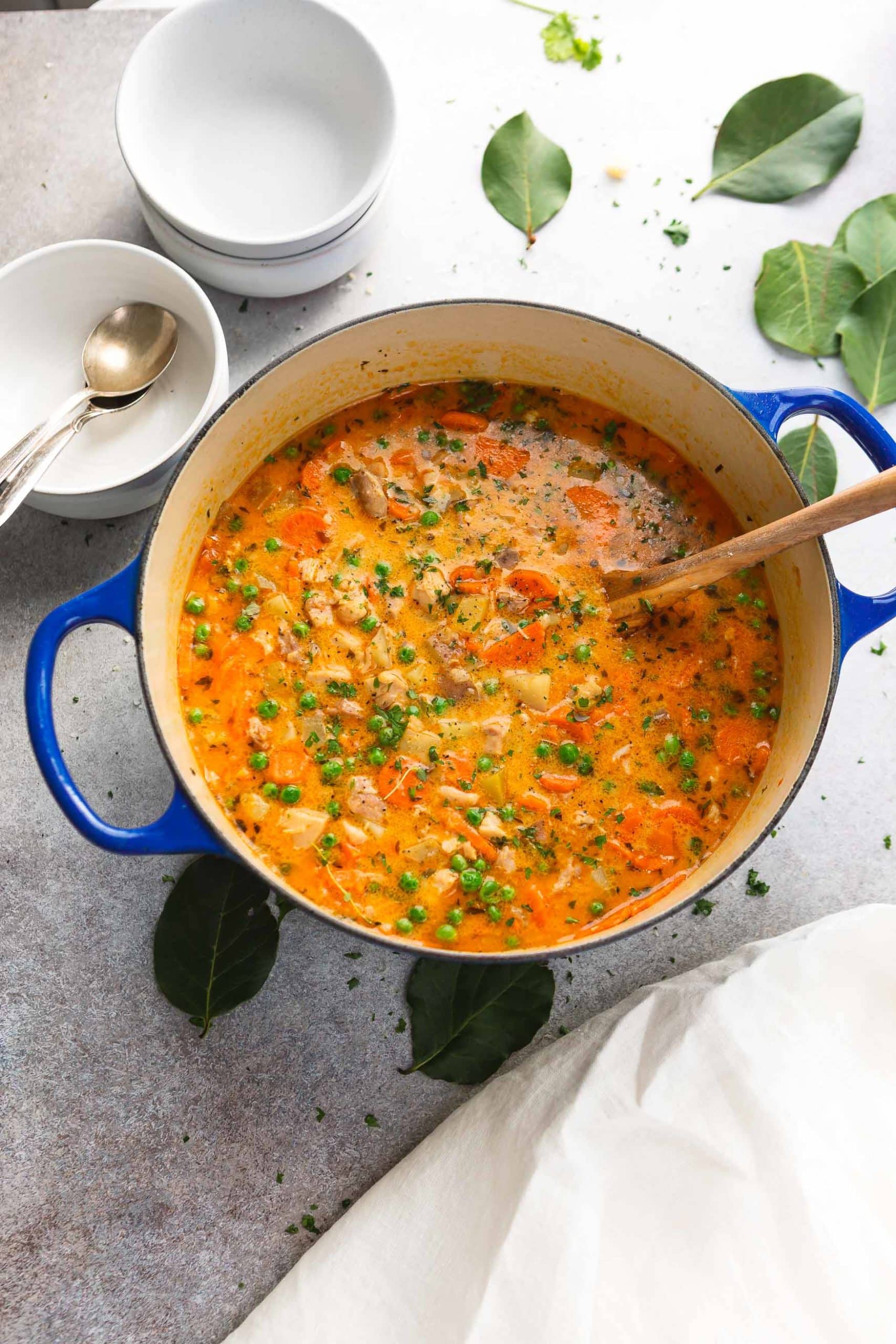 A pot of slow cooker chicken stew with a wooden spoon. The stew chicken and vegetables are in a thick yellow-red gravy and there are bright green herbs with the vegetables. A stack of bowls and spoons are in the background.