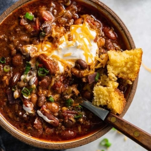 A bowl of chili. The chili has ground beef, beans and a rich sauce. It is topped with sour cream and shredded cheese and a piece of cornbread is on the edge of the bowl.