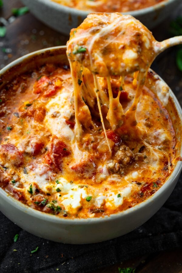 a photo of a bowl of lasagna soup topped with melted cheese and someone pulling a spoon out of the bowl with cheese stringing from it. a photo of a bowl of lasagna soup topped with melted cheese and someone pulling a spoon out of the bowl with cheese stringing from it.