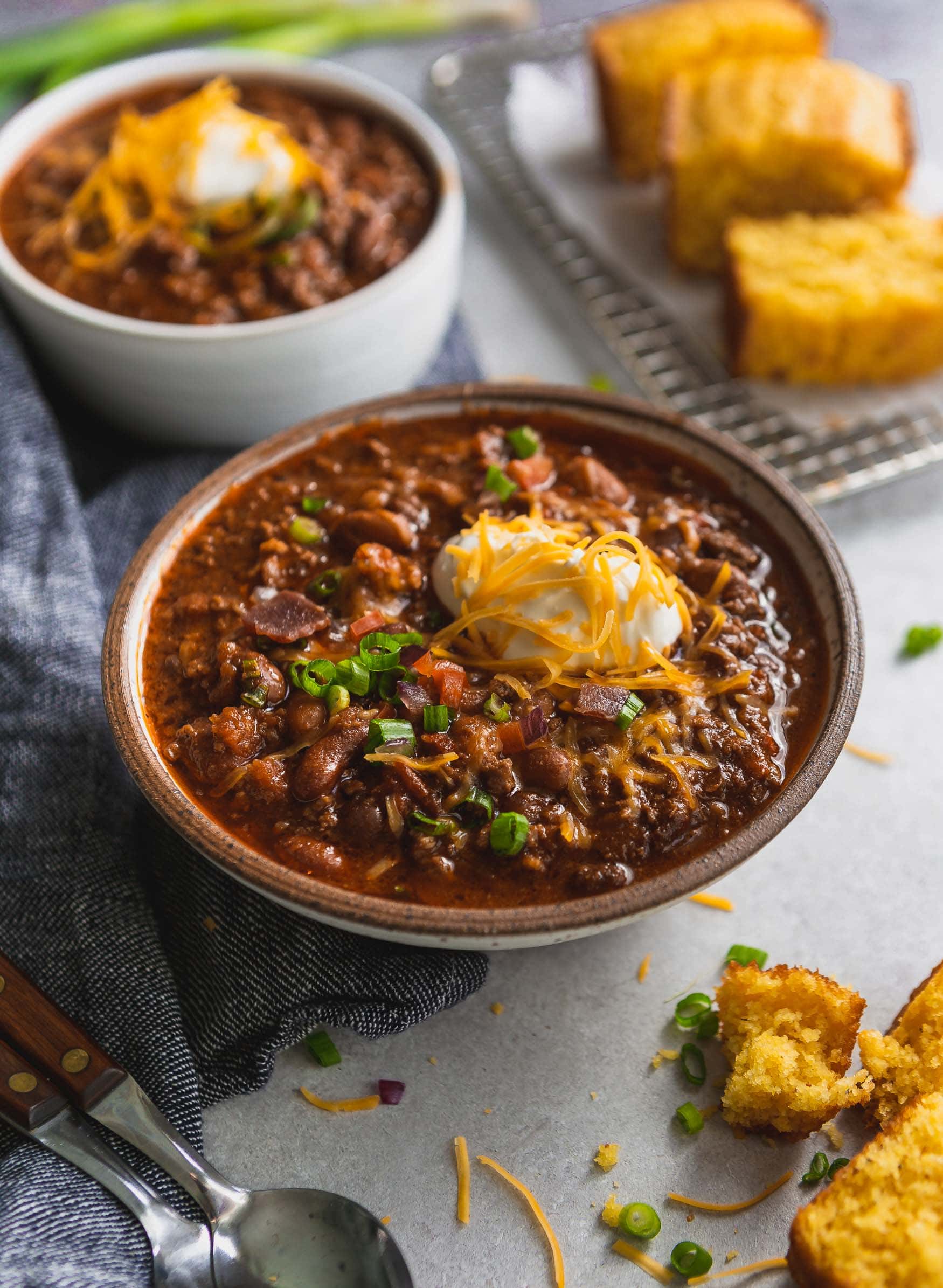 A large bowl of the best chili topped with sour cream and grated cheese. Another bowl for chili is in the background and there are a few pieces of cornbread next to the chili.