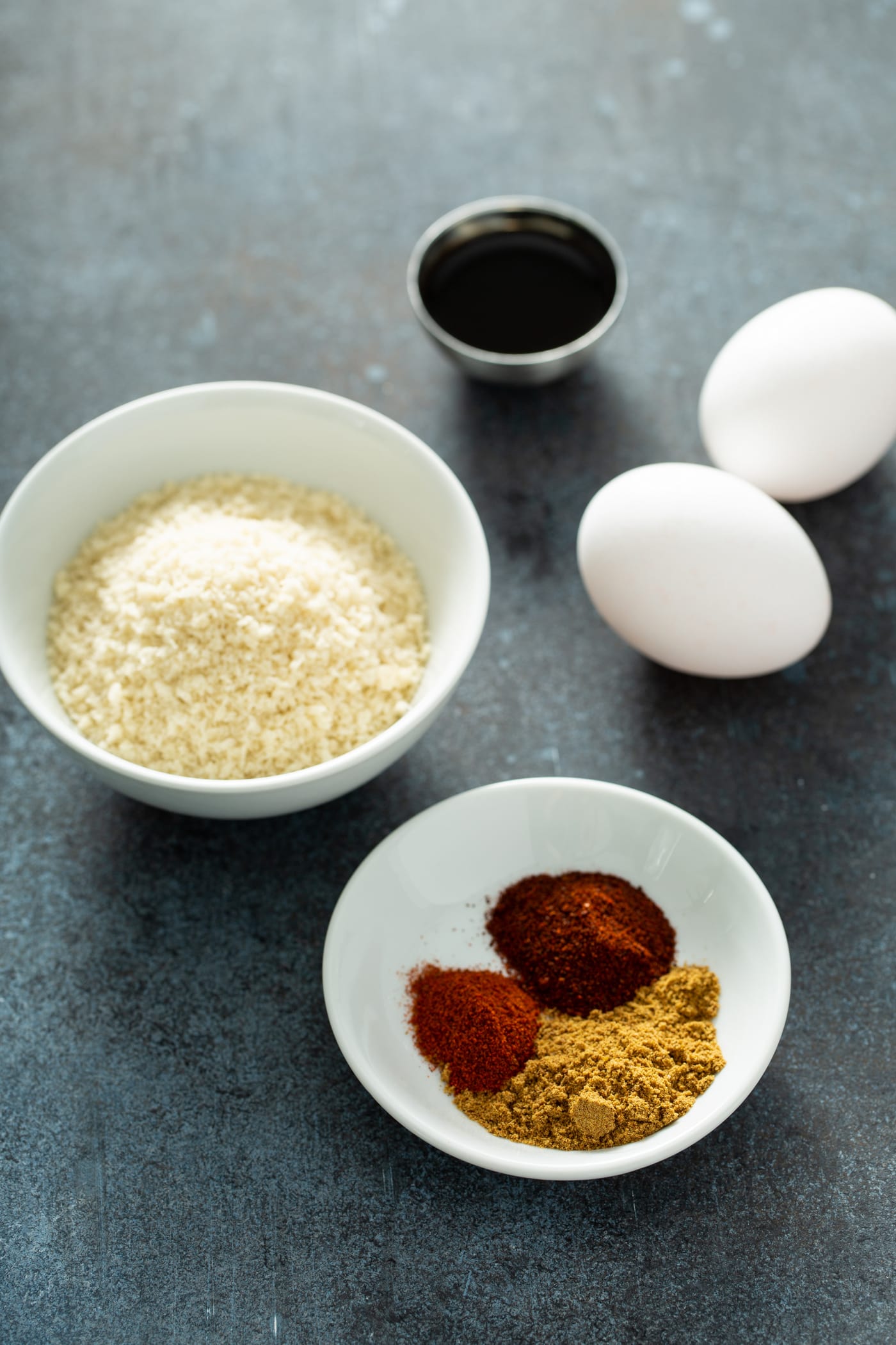 A white bowl with bread crumbs, a white bowl with cumin, paprika and chili powder, two white eggs, and a small container of Worcestershire Sauce. 