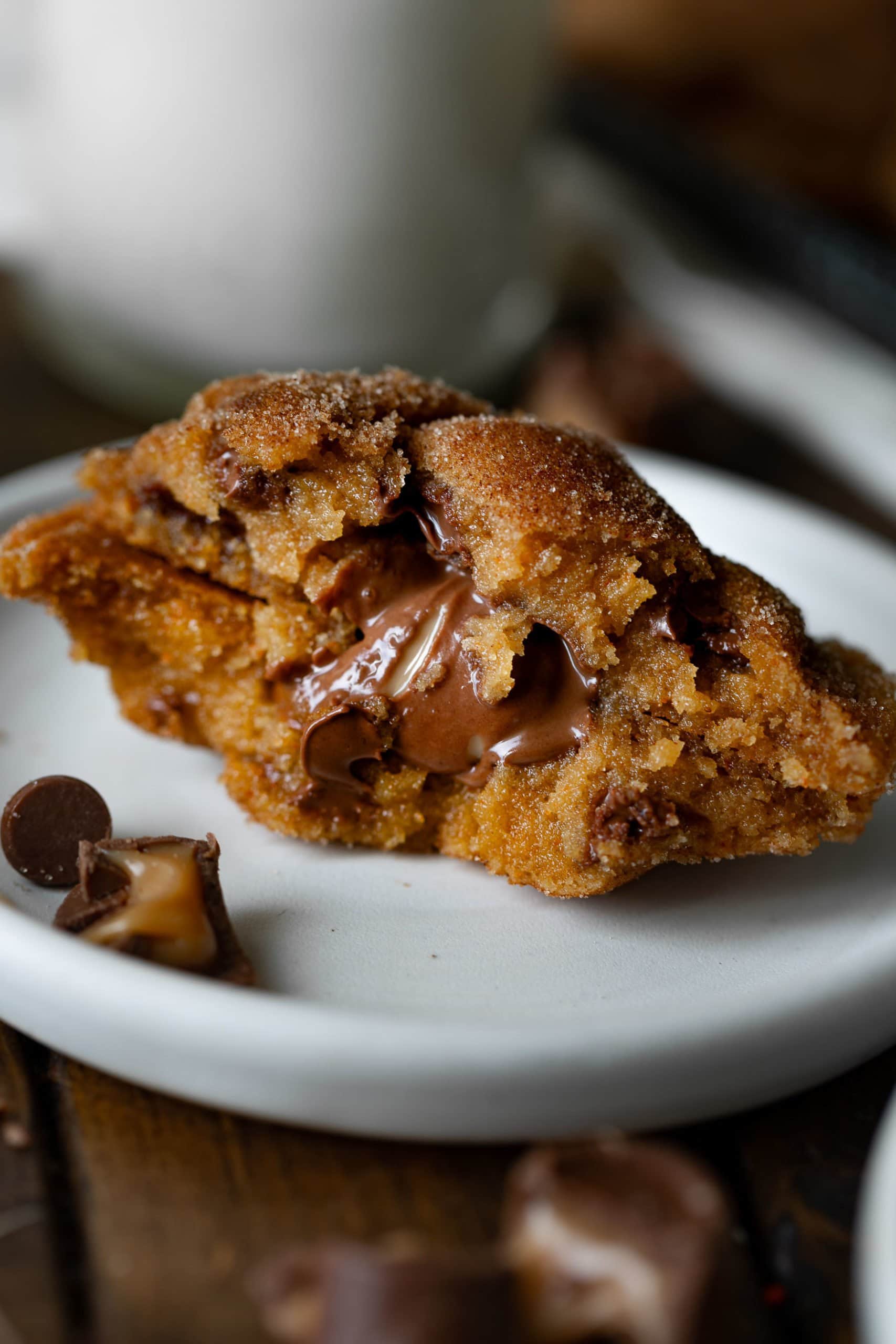 a photo of a Rolo stuffed pumpin pudding cookie sitting on a white dessert plate. The cookie has been broken in half with the two sides stacked on top of each other so you can see the gooey center.