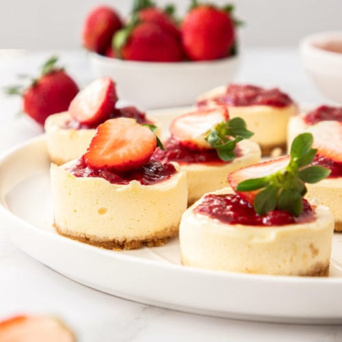 A large white plate with a batch of mini strawberry cheesecakes on it