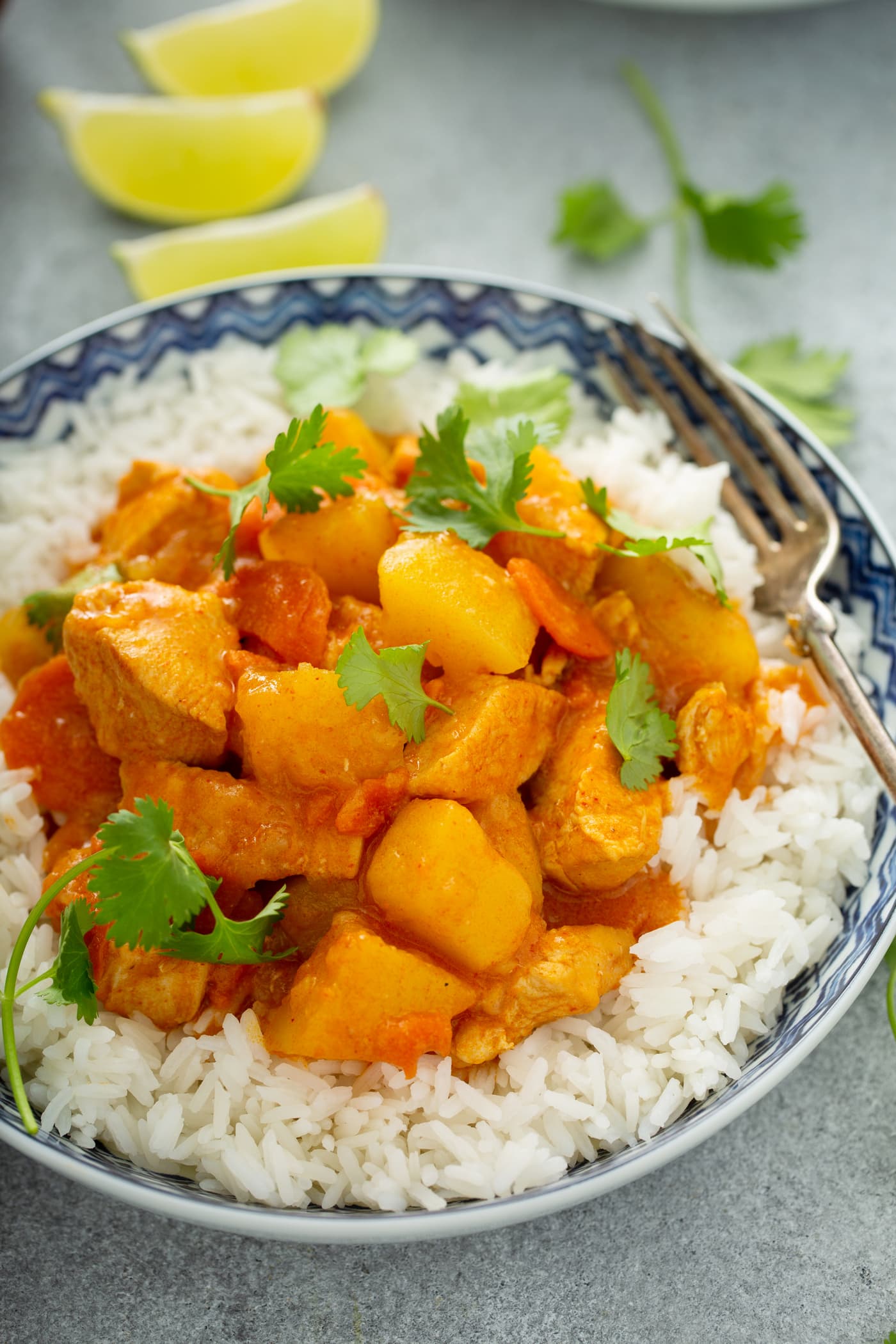 A bowl of white rice with chicken curry on top. there are chunks of chicken, carrots, and potatoes in a curry sauce. The curry is sprinkled with fresh parsley.