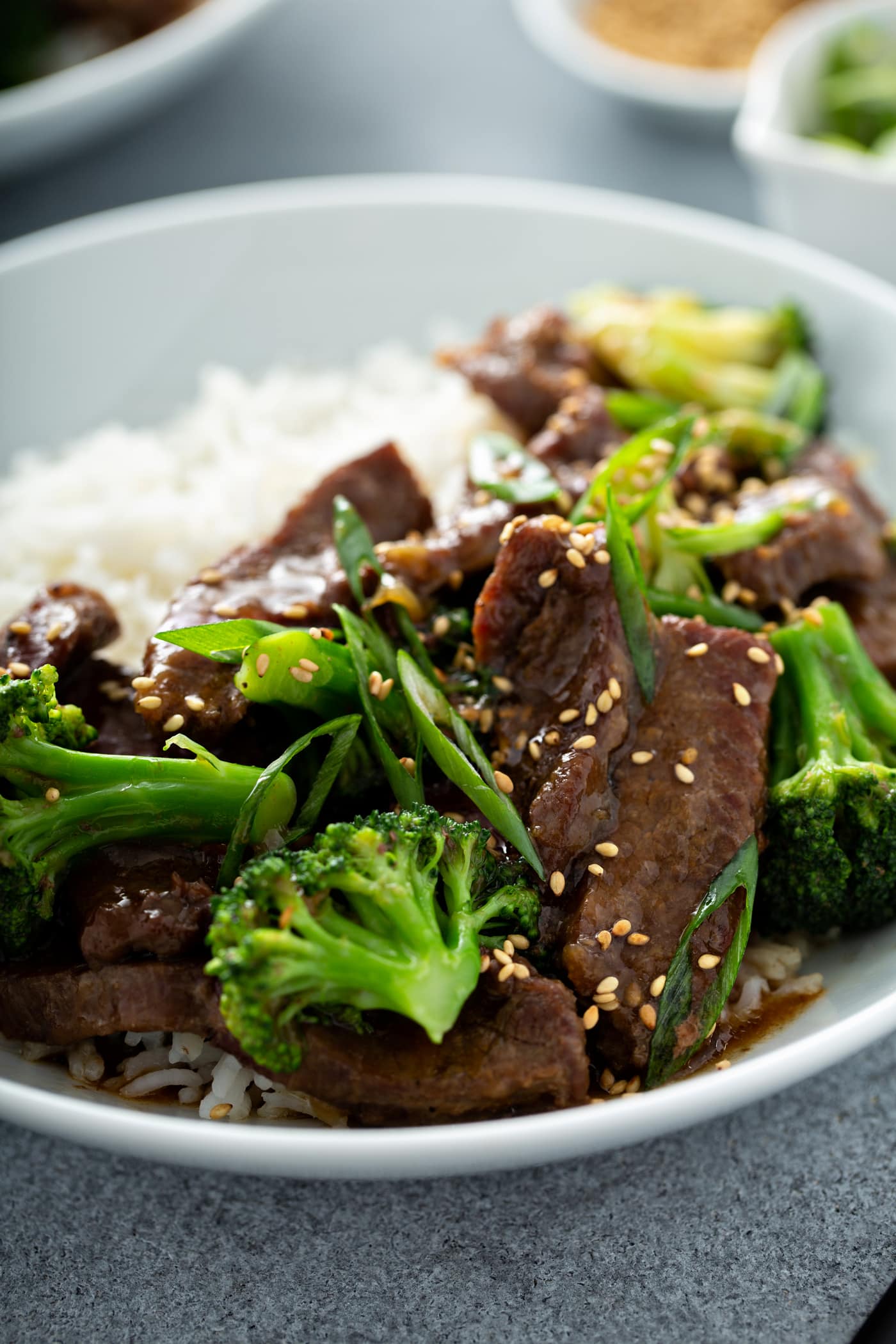A photo of a white bowl of beef and broccoli garnished with toasted sesame seeds and green onions.