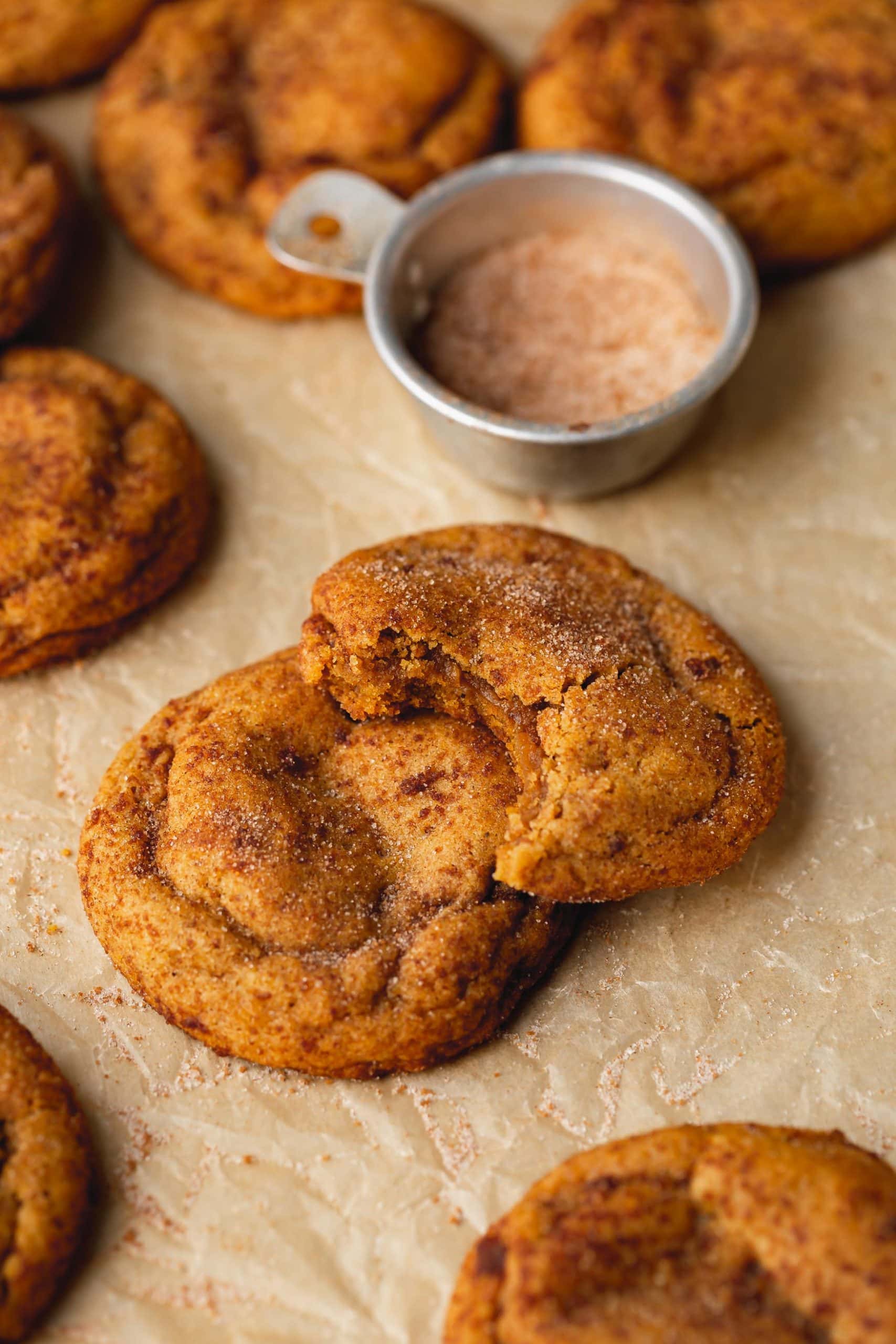 Two chewy pumpkin cookies stacked on top of each other. The cookie on top has a bite taken out of it and there is another cookie and a container of cinnamon sugar in the background.