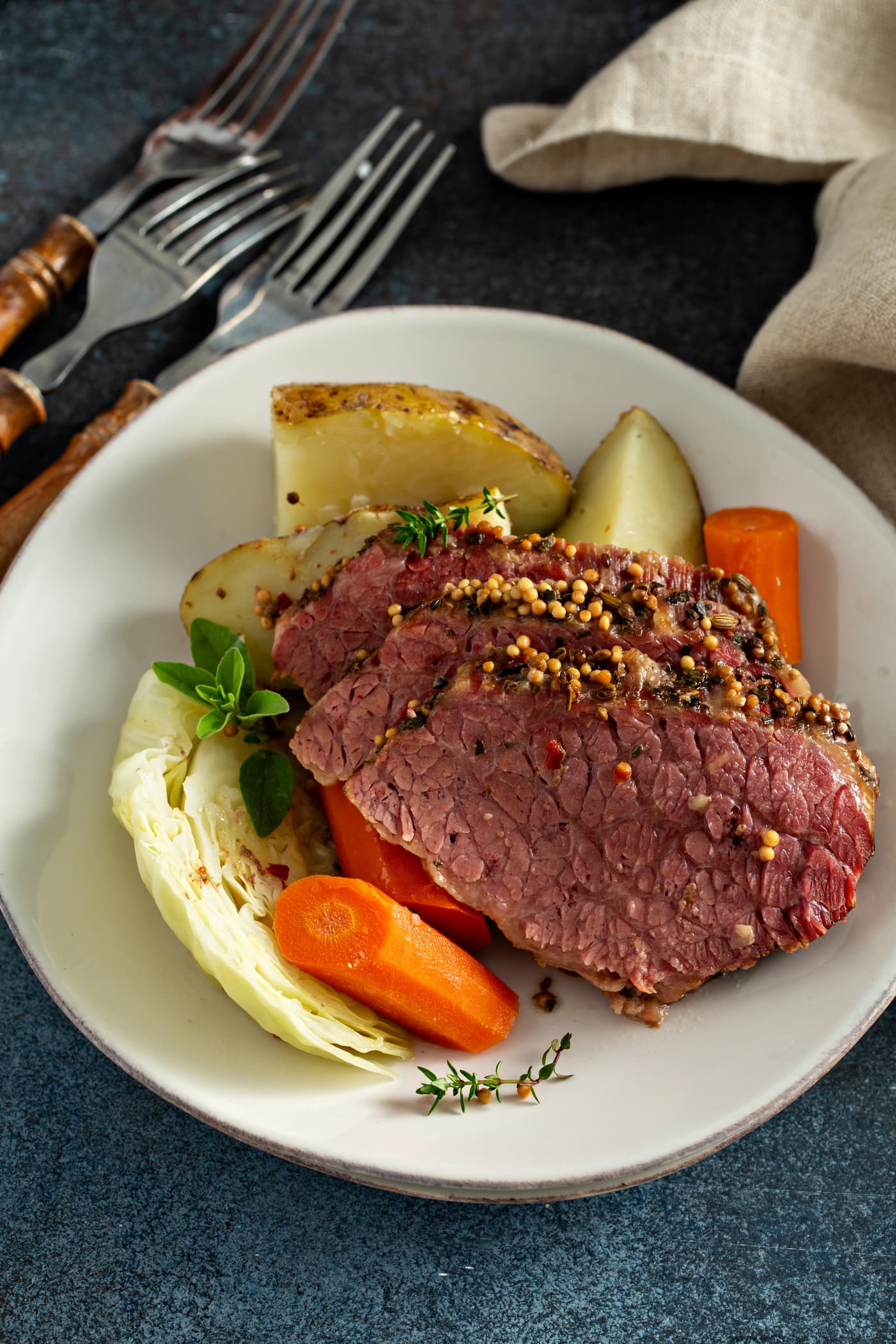 Three slices of corned beef resting on potatoes and carrots and cabbage on a white serving plate. There are forks next to the plate.