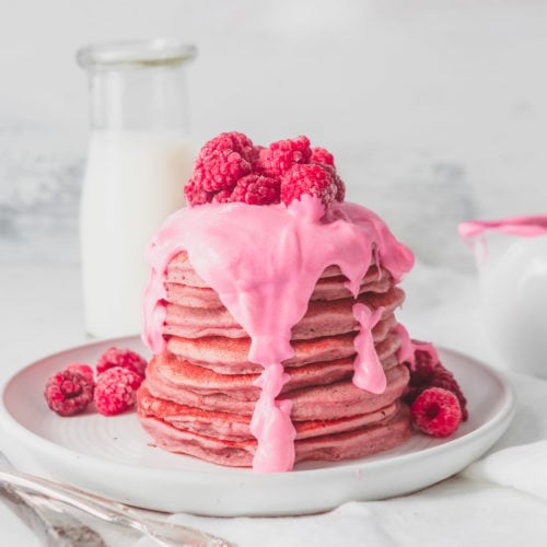 A large stack of pink pitaya pancakes with a thick pink chocolate sauce poured over the top. There are fresh raspberries on top of the sauce and raspberries on the plate around the pancakes.