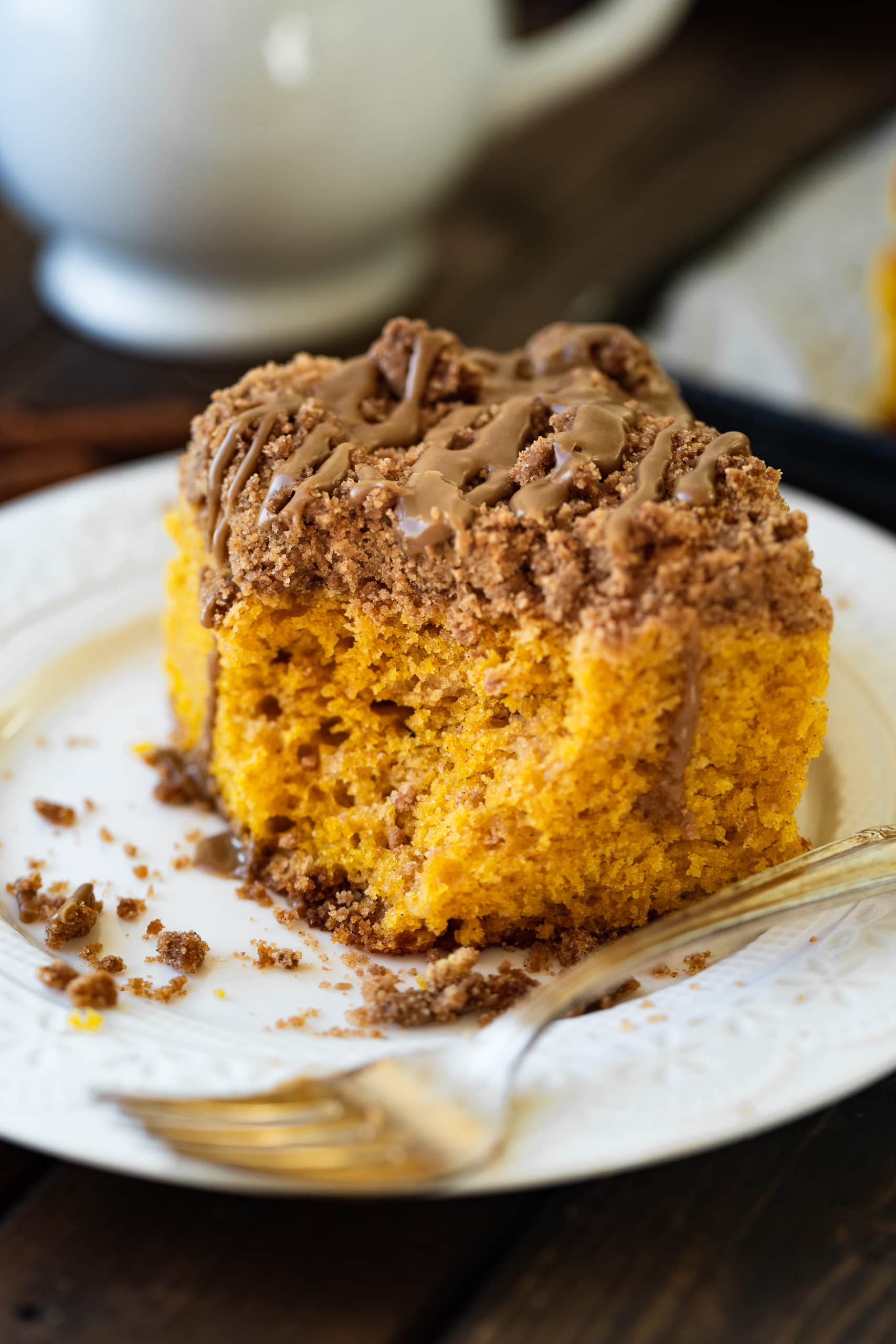 a photo of a piece of pumpkin coffee cake topped with cinnamon streusel and maple glaze sitting on a white dessert and bite taken out of the corner.