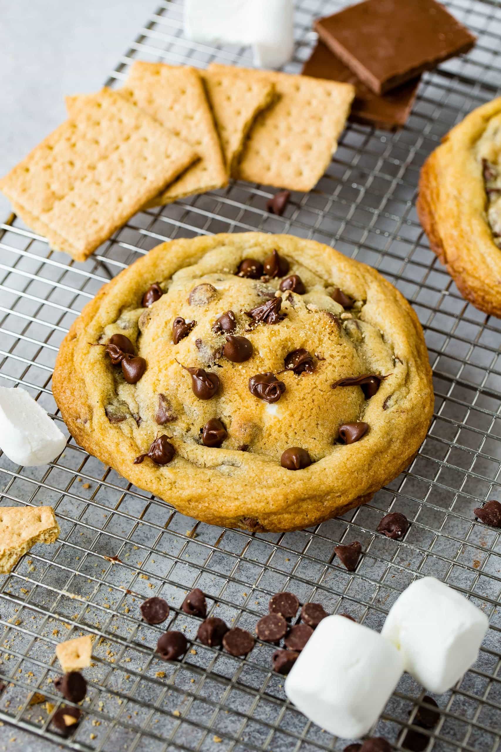 A s'mores stuffed chocolate chip cookie. There are graham crackers, chocolate chips and marshmallows in the background.