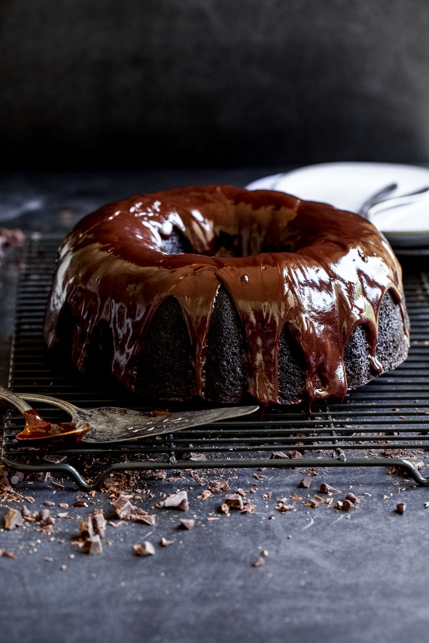 A zucchini chocolate bundt cake with a rich chocolate glaze. The cake is on a wire cooling rack and has mini chocolate chips scattered under and around the rack.