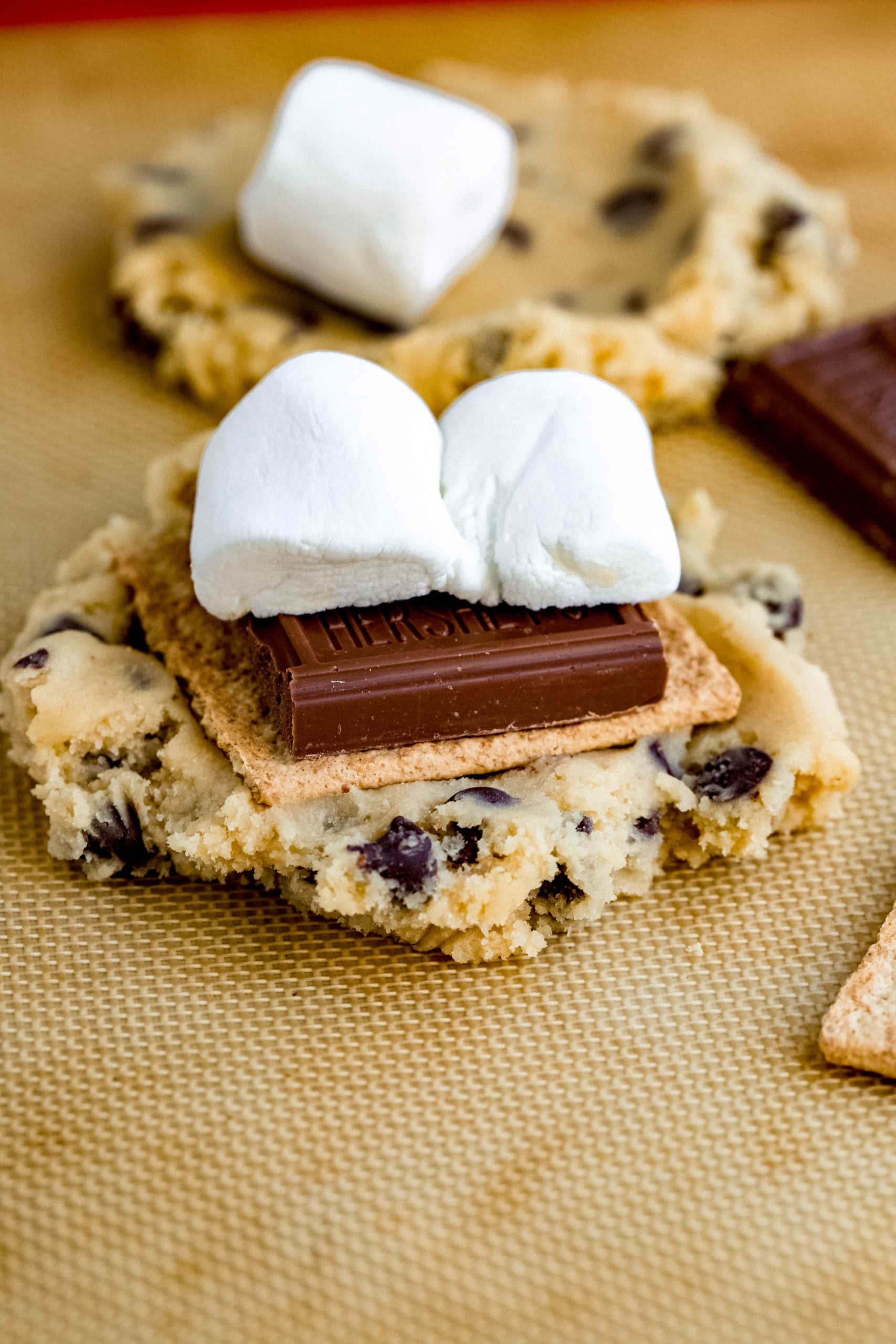 A baked chocolate chip cookie with a graham cracker, chocolate bar, and marshmallow on top. Another cookie and marshmallow are in the background.