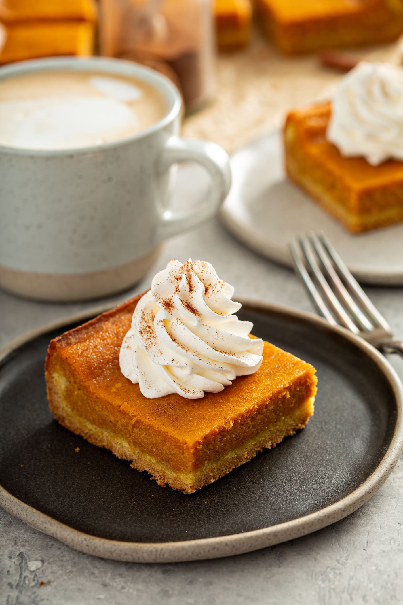 A piece of pumpkin gooey butter cake topped with whipped cream and sprinkled with cinnamon, sitting on a dessert plate. A fork lays beside the plate and another piece of cake with whipped cream and a white mug are in the background.