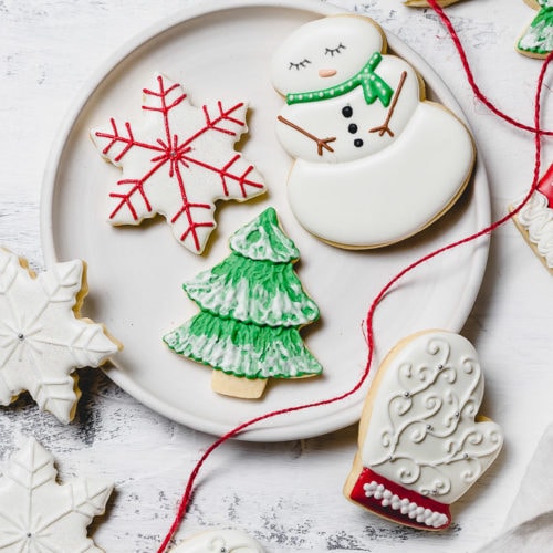 A plate with Christmas sugar cookies decorated with royal icing. There is a snowman with a green scarf, a white mitten with red trim, a green tree with white snow accents, and a white star with red accents. A red ribbon is draped over the edge of the plate.