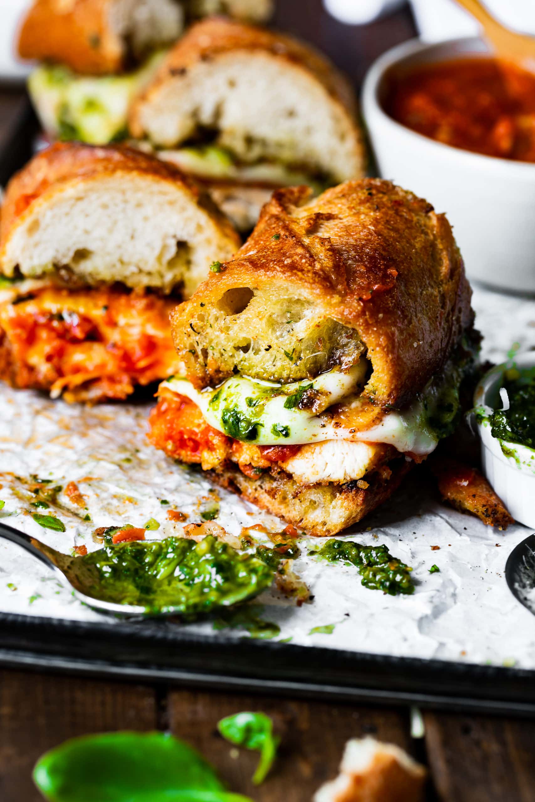 a photo of a chicken parm sandwich that has been cut in half so you can see the cross section with a spoonful of pesto sitting in front of it.