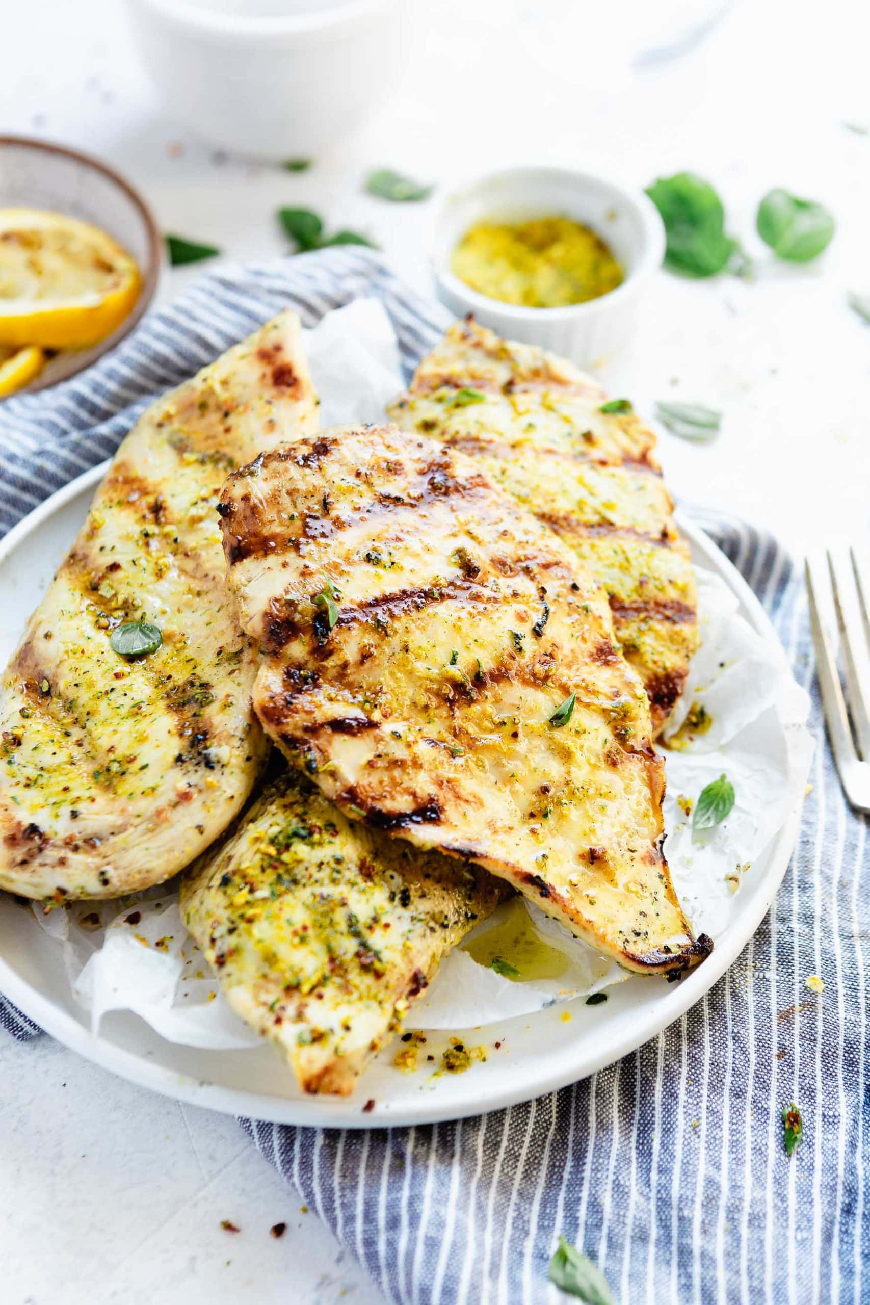 A plate of grilled chicken breasts. There are grilled lemon slices in the background and a knife and fork next to the plate of chicken. 