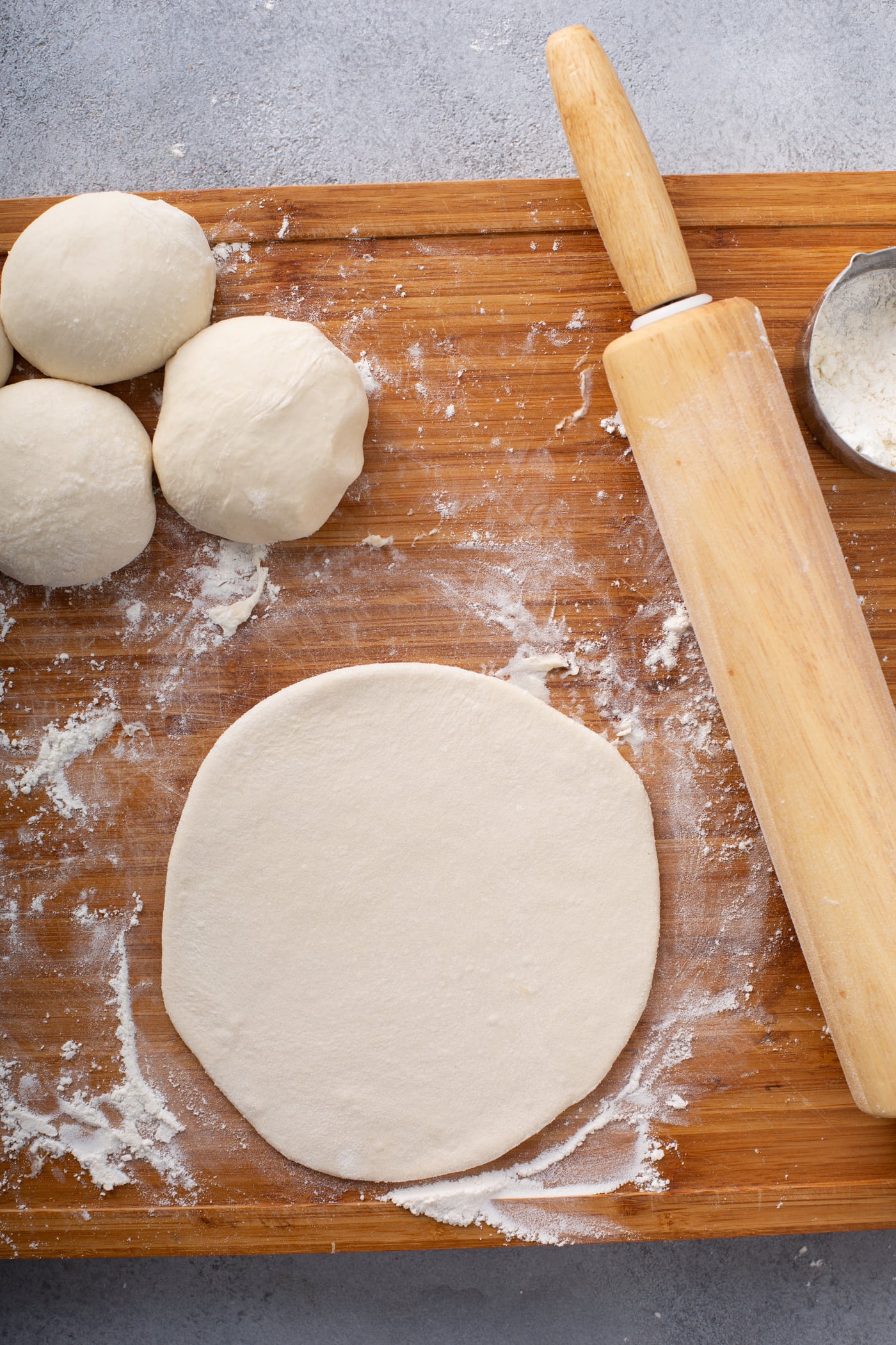 A photo of a portion of dough rolled out into a flat circle on a floured wooden cutting board with a wooden rolling pin sitting next to it and unrolled portions of dough also sitting next to it.