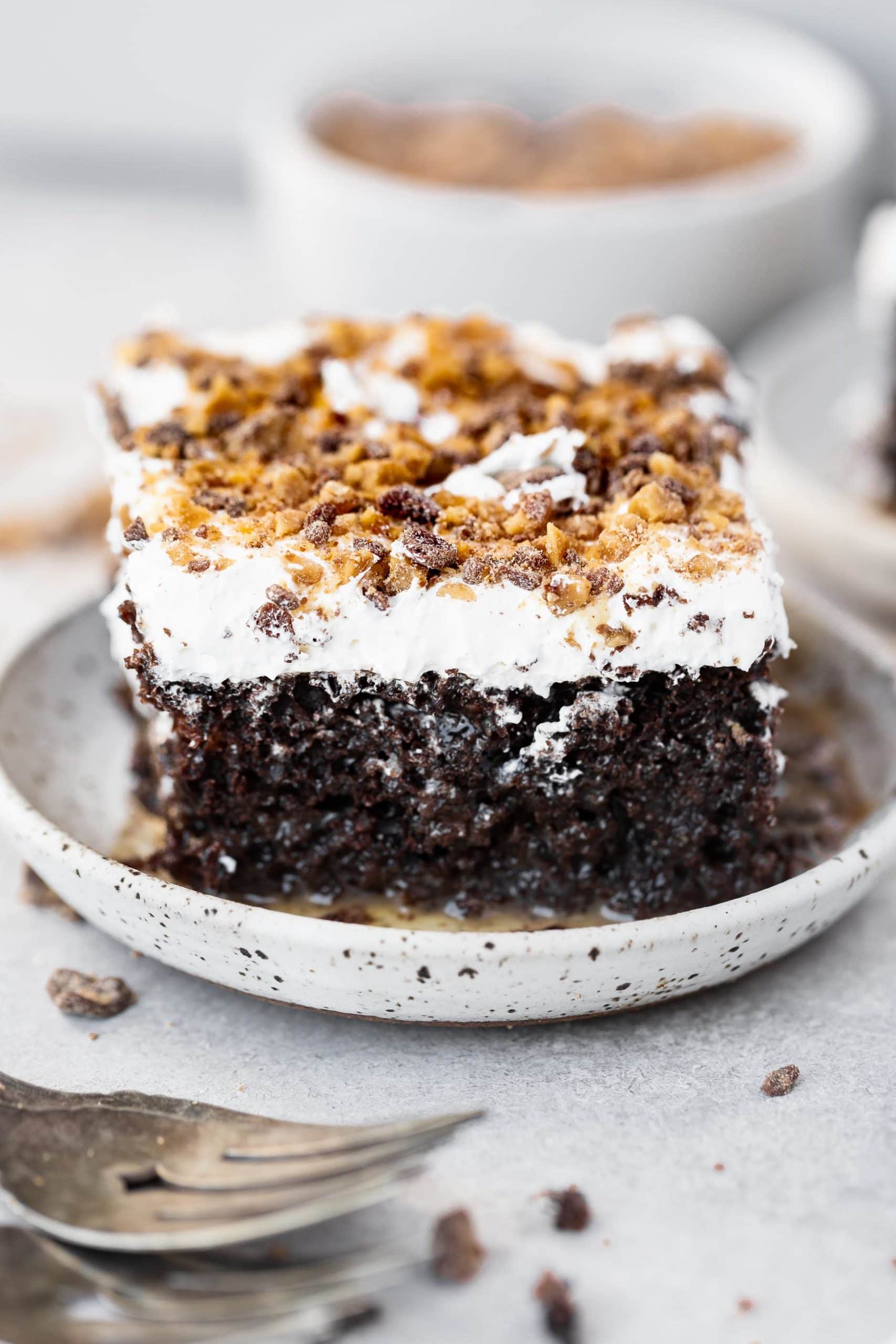 a photo of a single piece of chocolate cake topped with whipped cream and toffee bits and the cake is oozing caramel on the saucer it is sitting on.