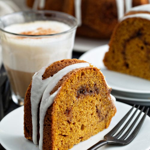 A slice of sour cream cinnamon swirl pumpkin bundt cake on a dessert plate with a fork on the plate next to the cake. The cake has a drizzle of white glaze on top. another slice of cake on a dessert plate, and the rest of the bundt cake on a cake platter are in the background.