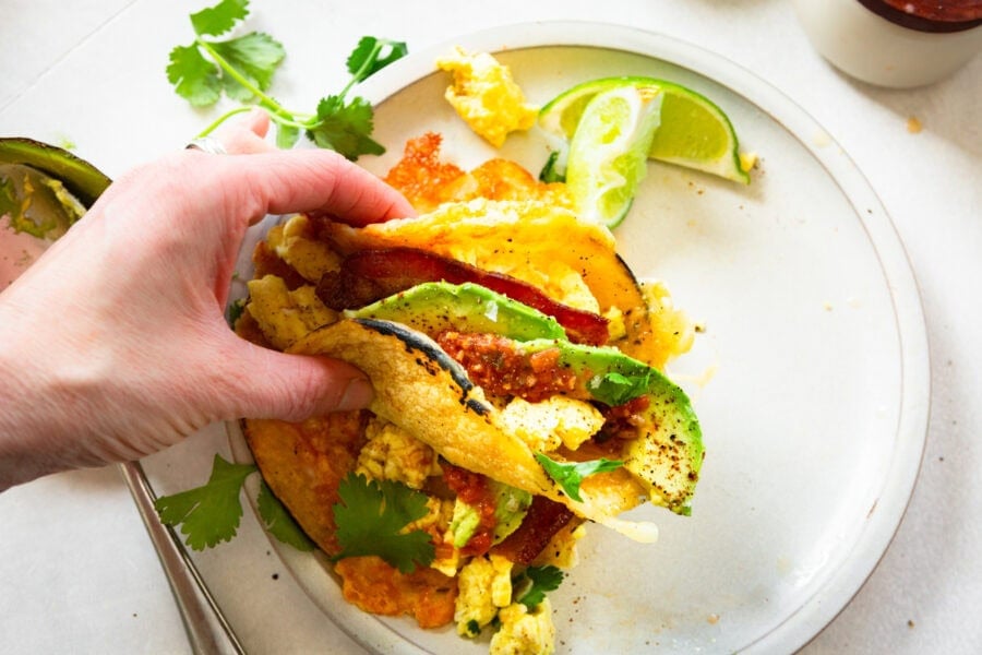 a hand holding a breakfast taco with eggs, bacon, cheese and avocado slices loaded in a corn tortilla